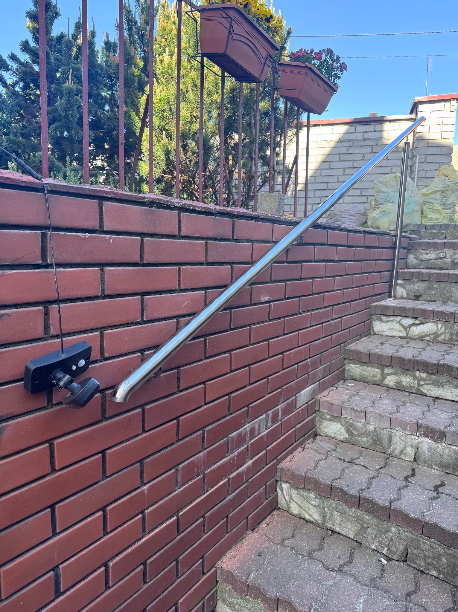 Nierdzewna poręcz zamontowana na ceglanym murze przy schodach zewnętrznych. Na murze widoczna lampa. W tle roślinność i worki z materiałami budowlanymi.