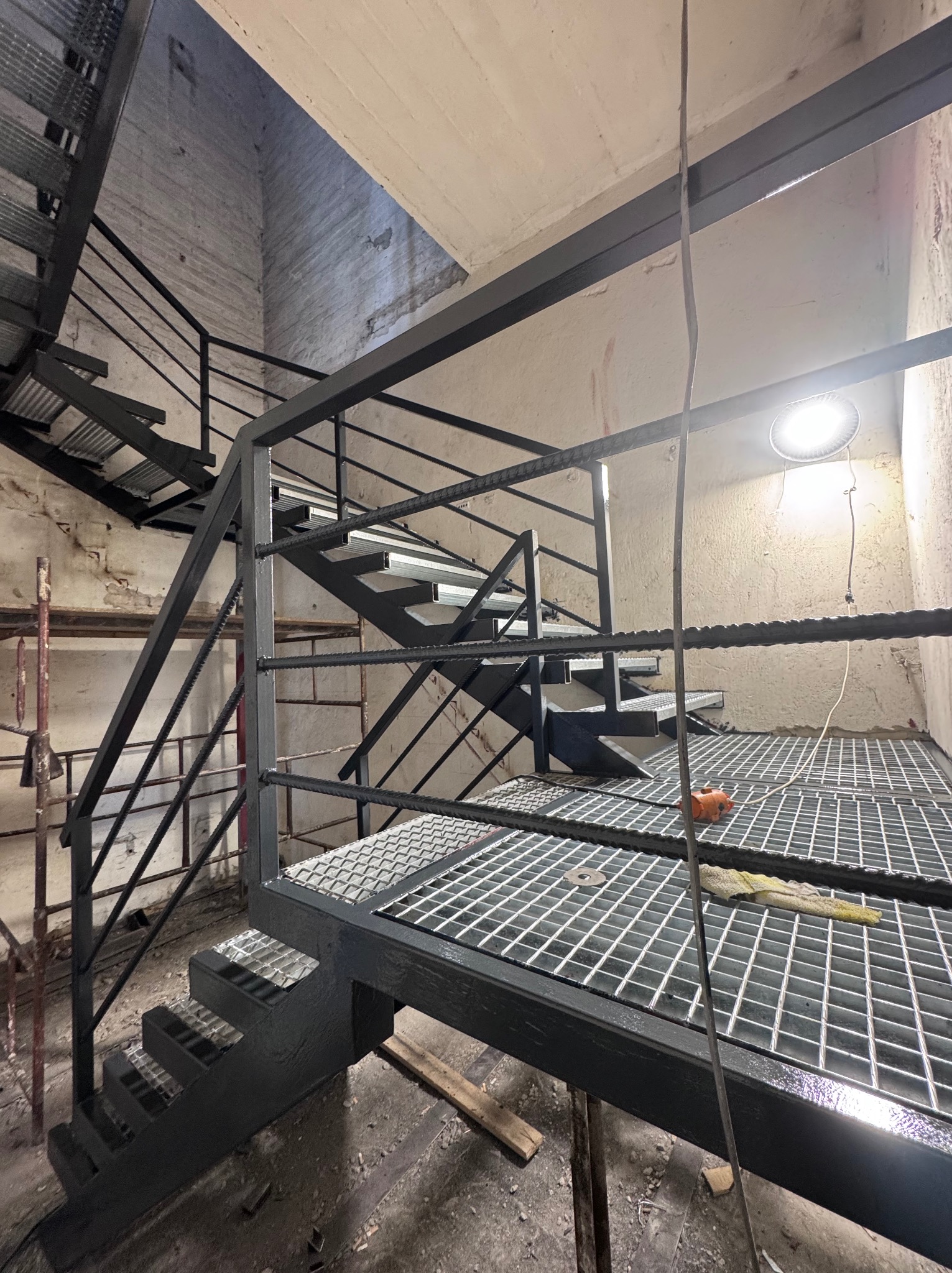 Metalowe schody z platformą kratową w surowym wnętrzu. Widoczne spawy i elementy konstrukcyjne, oświetlenie lampą. Kąt ujęcia od dołu.