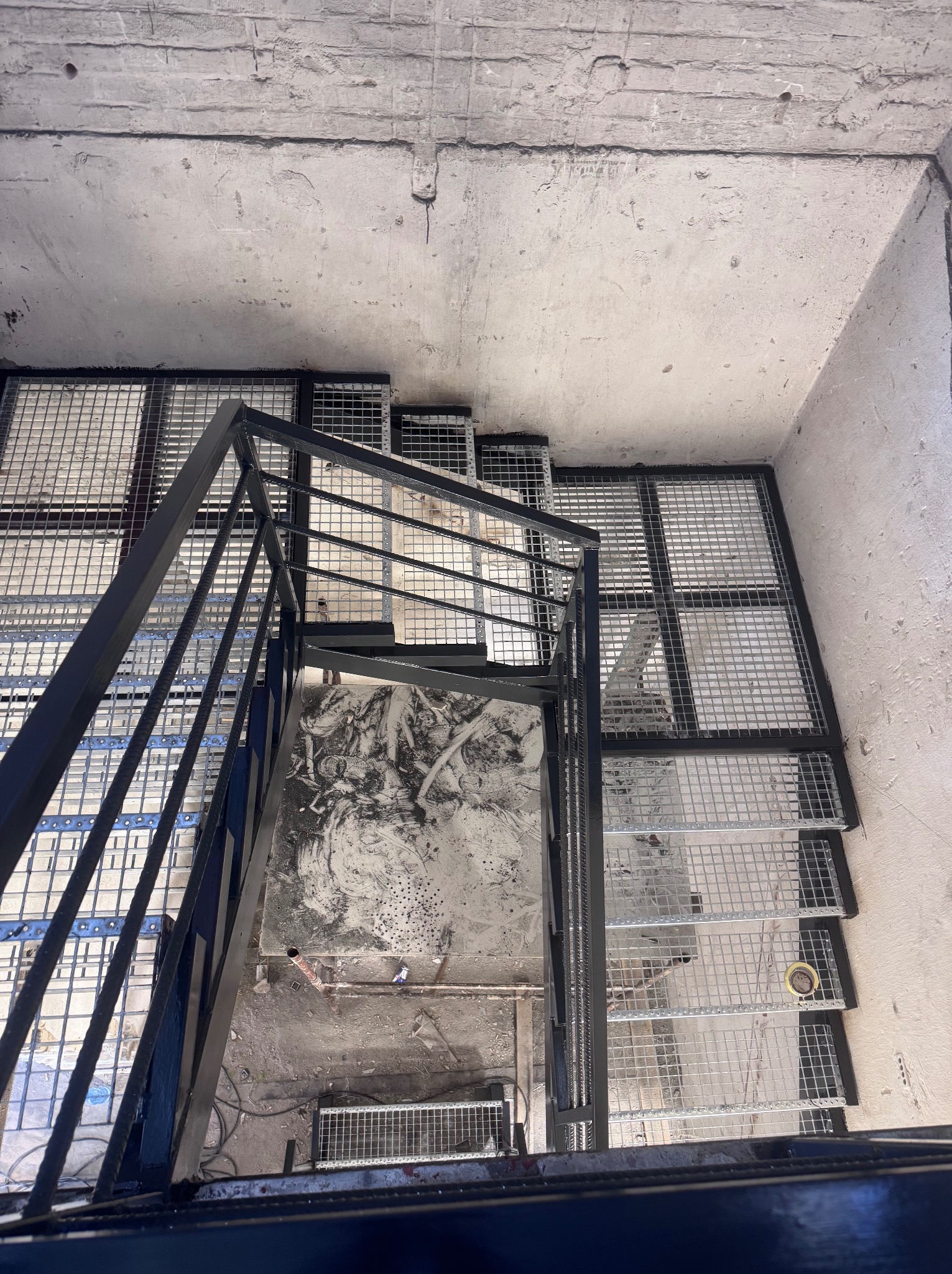 Metalowe schody z poręczą, widziane z góry, z siatkowymi stopniami w surowym wnętrzu. Widoczne ślady prac budowlanych na podłodze.