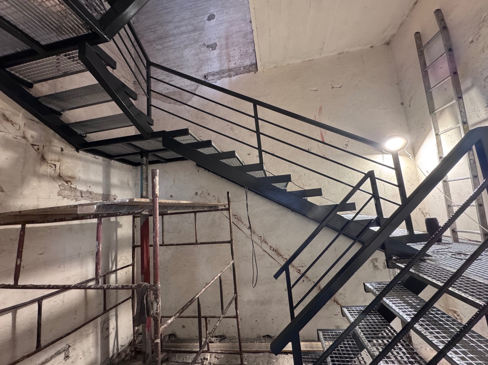 Metalowe schody z czarną balustradą w surowym wnętrzu, widoczne rusztowania, lampa oświetlająca. Industrialny styl i montaż schodów.