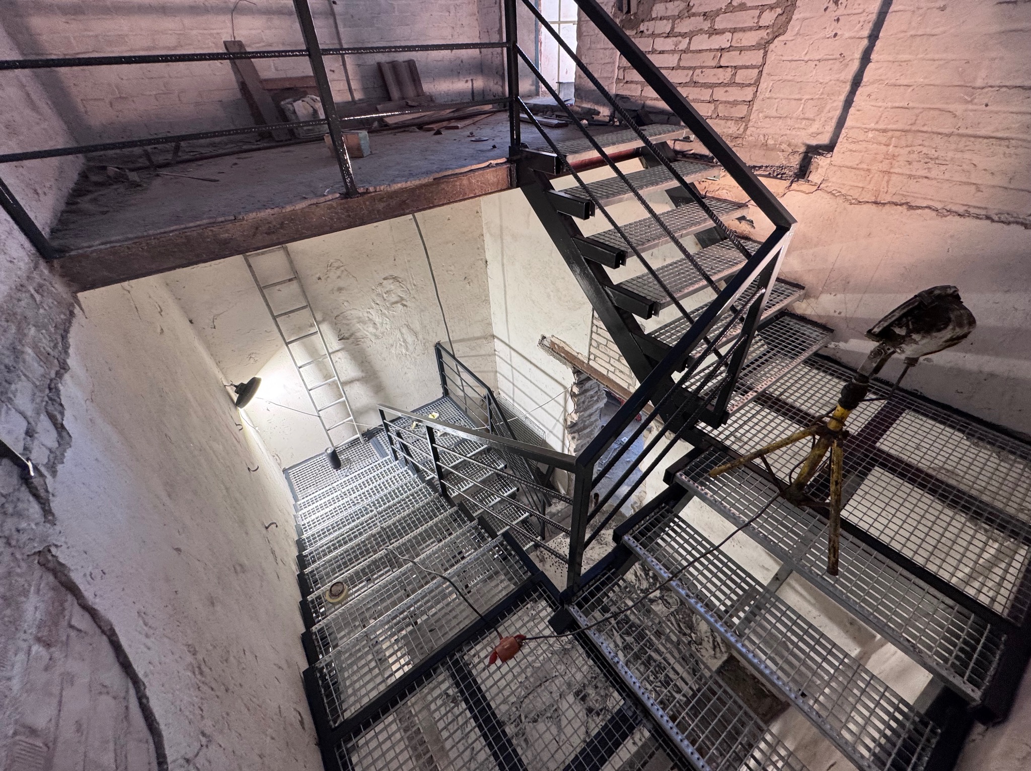 Metalowe schody kratowe z czarną poręczą w surowym wnętrzu, widok z góry. Na stopniach widoczne narzędzia i osprzęt. Fragment ceglanej ściany w tle.