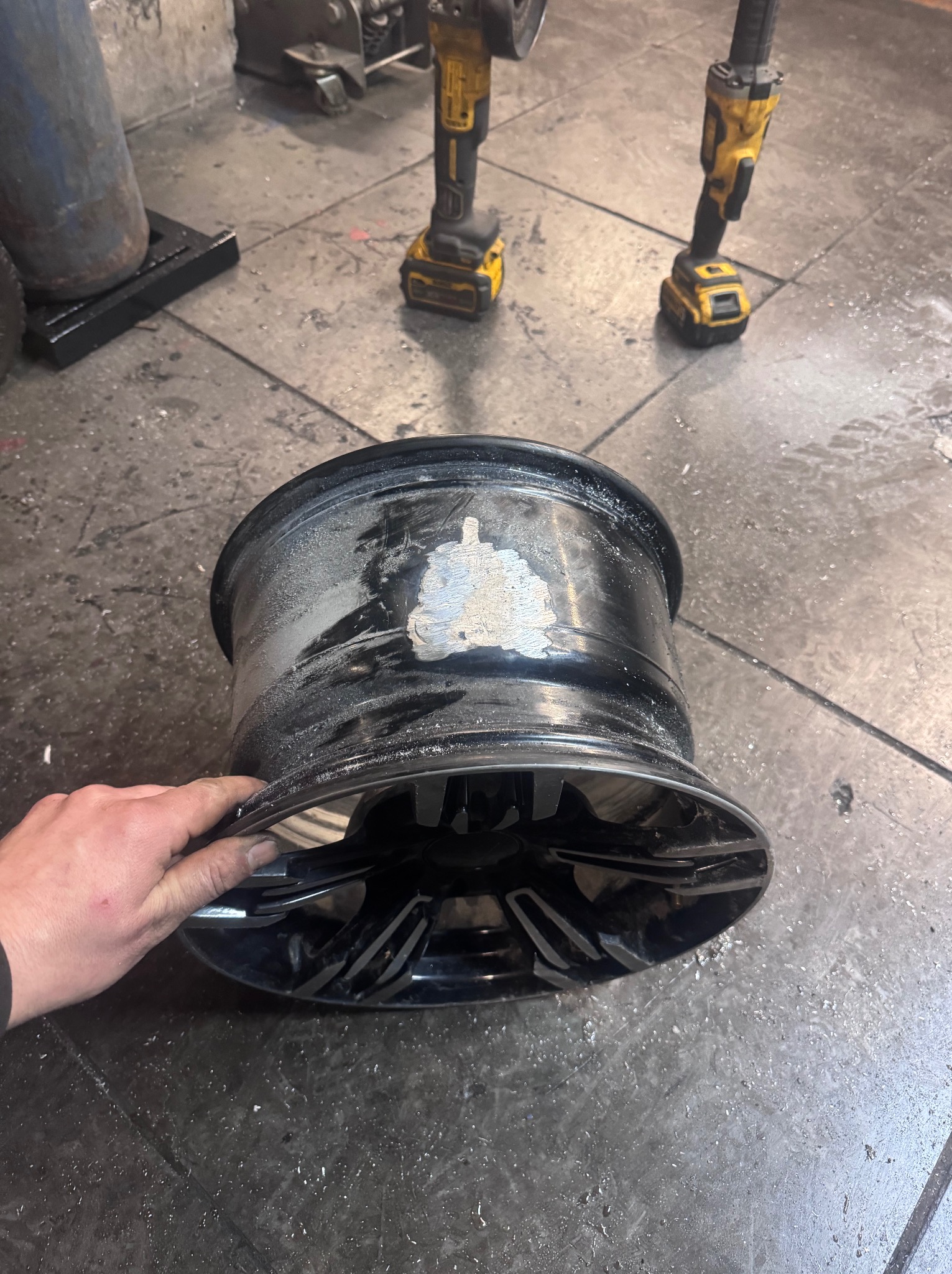 Czarna felga samochodowa w trakcie renowacji, z widocznymi śladami szlifowania i ręką osoby trzymającej felgę. W tle narzędzia na posadzce warsztatu.