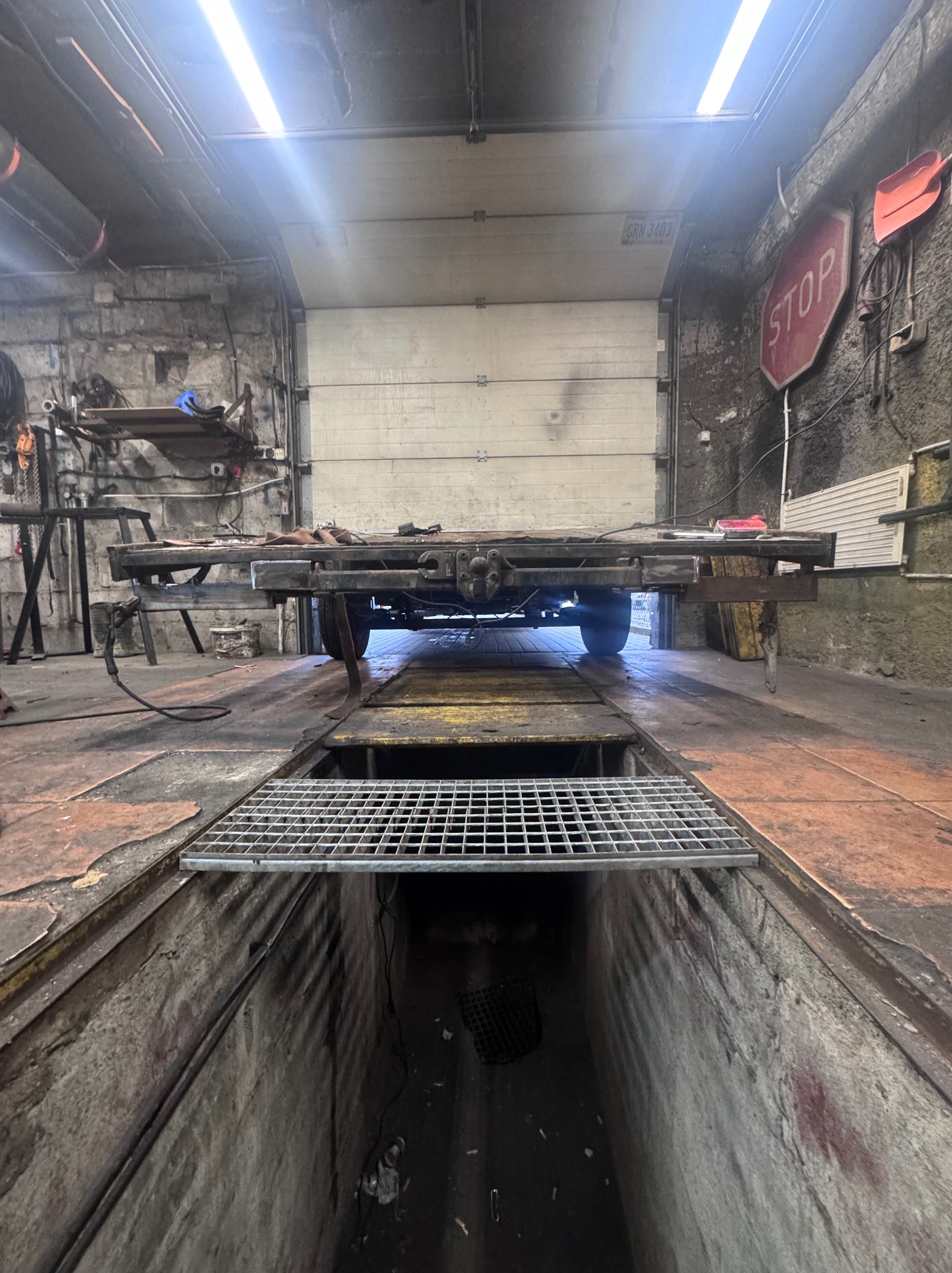 Warsztat ślusarski: rama przyczepy na kanale, spawarka i narzędzia w tle. Surowy, industrialny klimat.