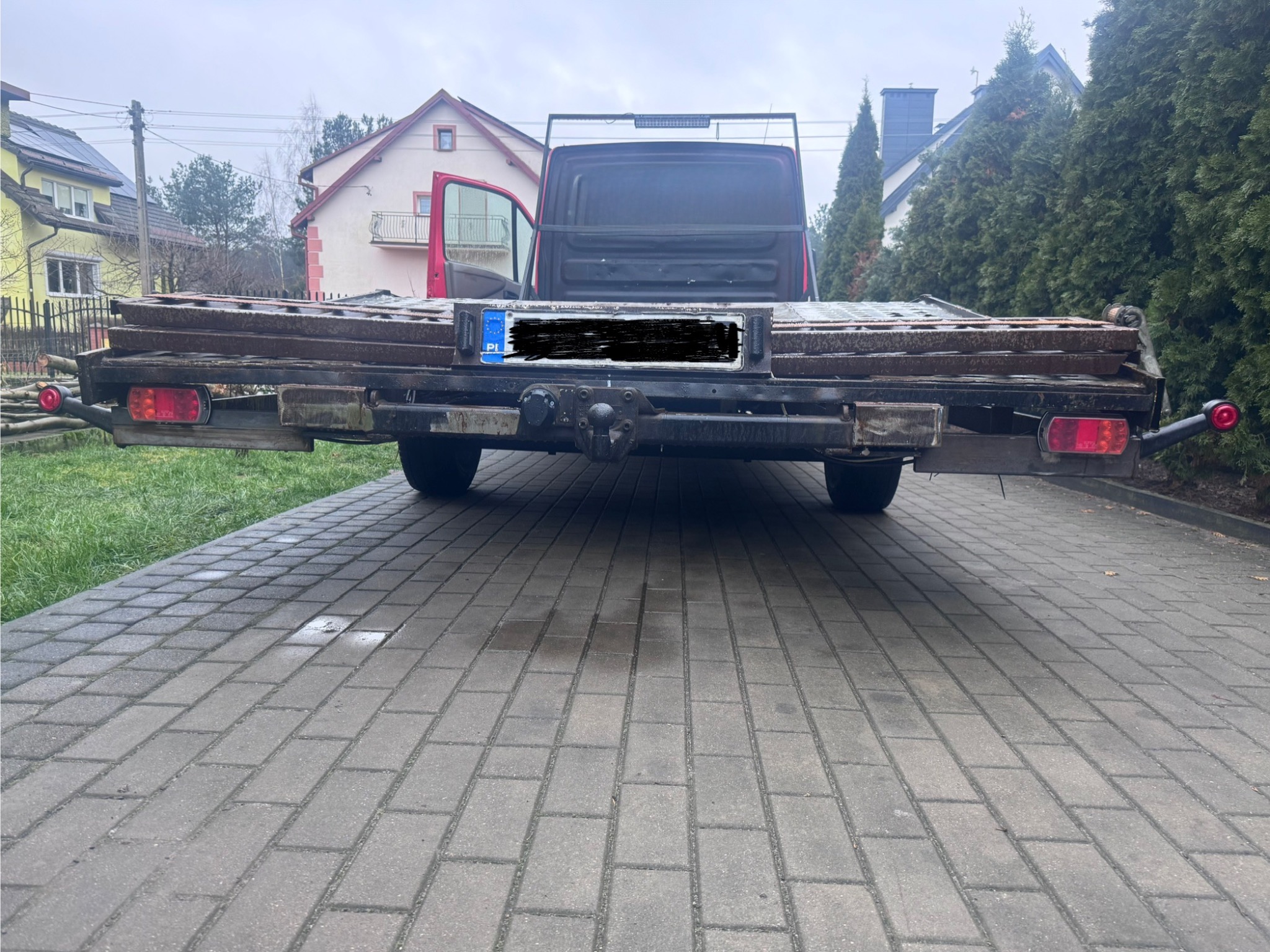 Widok z tyłu przyczepy z metalową konstrukcją, prawdopodobnie transportującej elementy metalowe. W tle domy jednorodzinne i kostka brukowa.