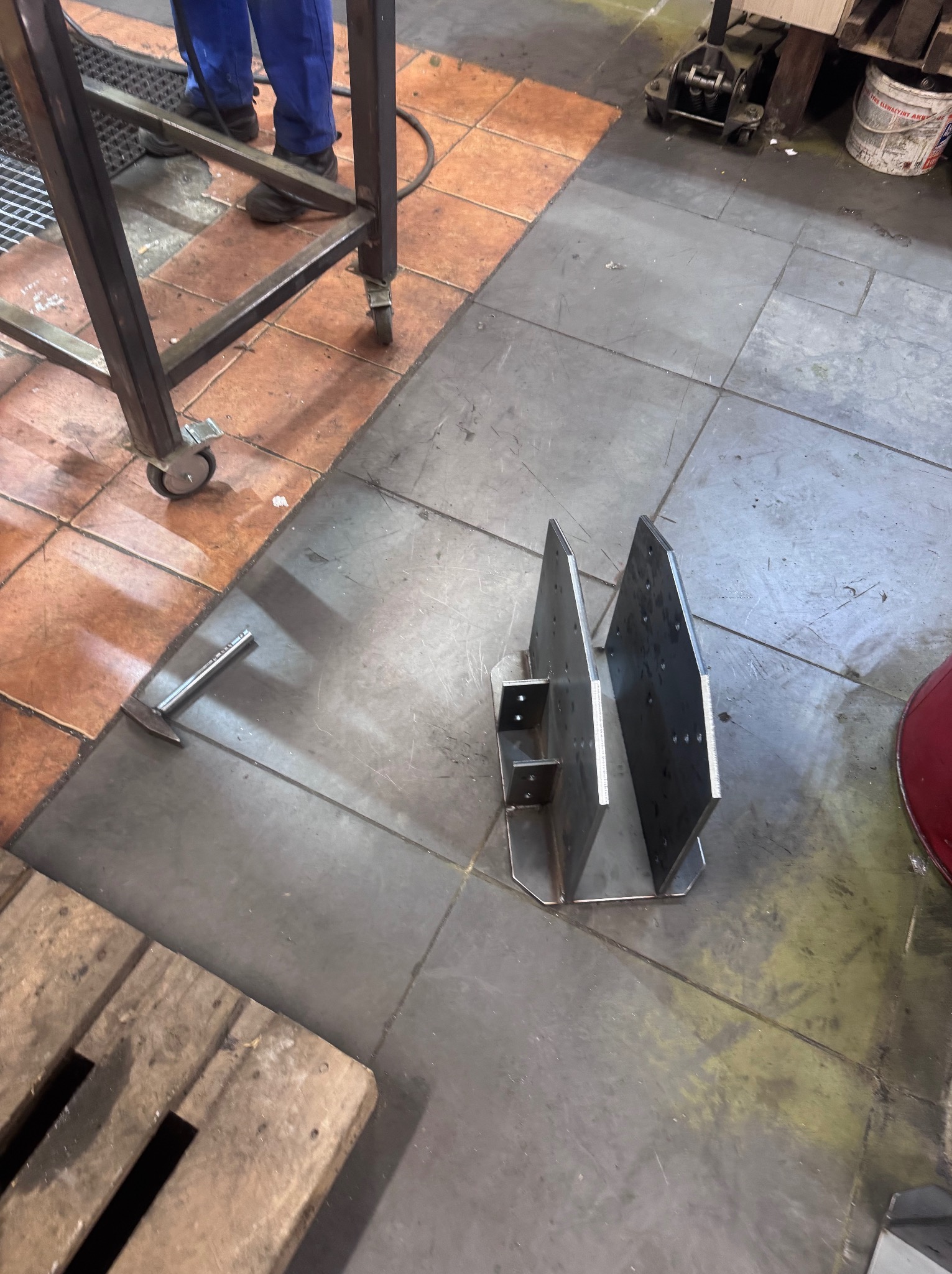 Metalowa konstrukcja spawana z dwóch pionowych płyt, leżąca na posadzce warsztatu, obok młotek i fragmenty stołu spawalniczego na kółkach.