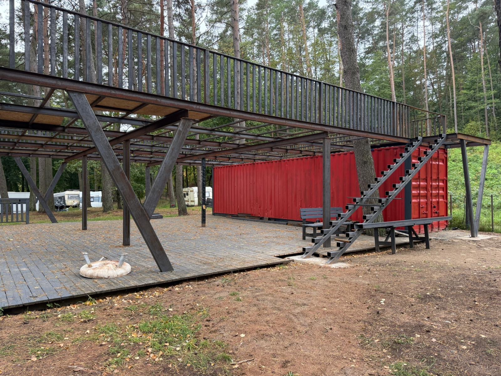 Metalowa konstrukcja z pomostem i schodami przy czerwonym kontenerze, otoczona drzewami. Nietypowe połączenie architektury i natury.