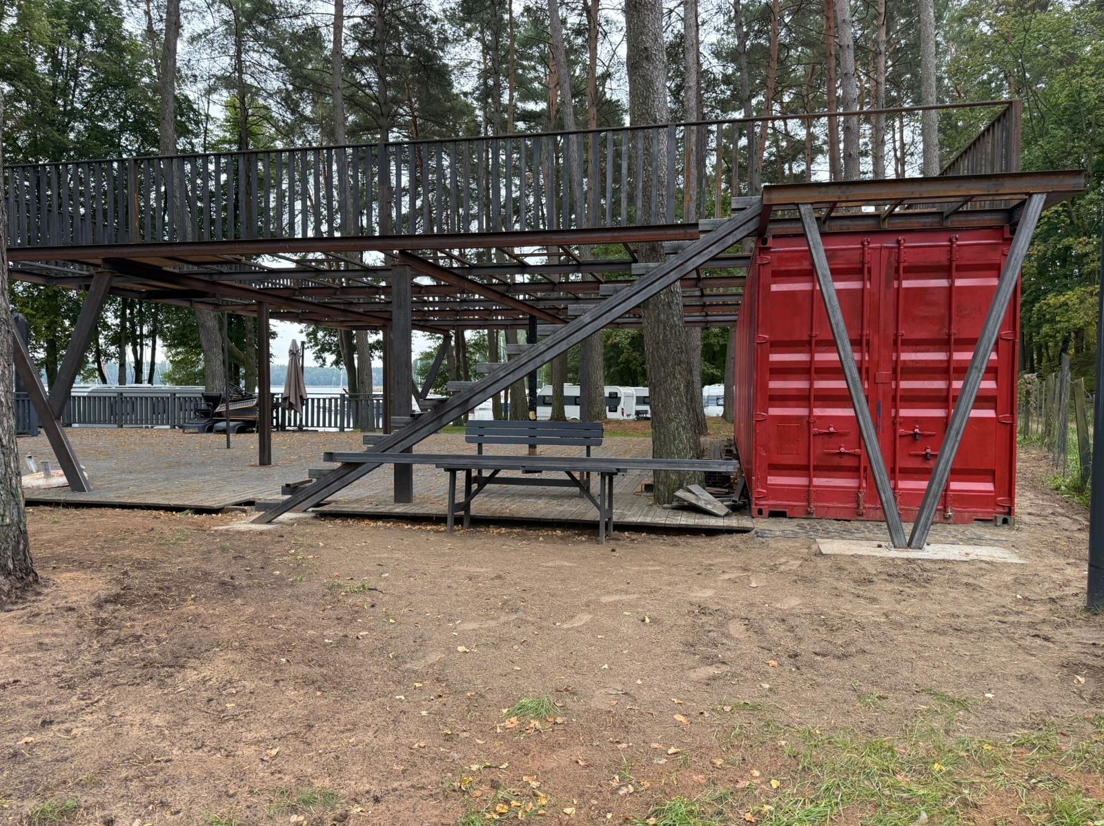 Metalowa konstrukcja z platformą widokową i schodami, połączona z czerwonym kontenerem, otoczona drzewami. Nietypowa architektura w plenerze.