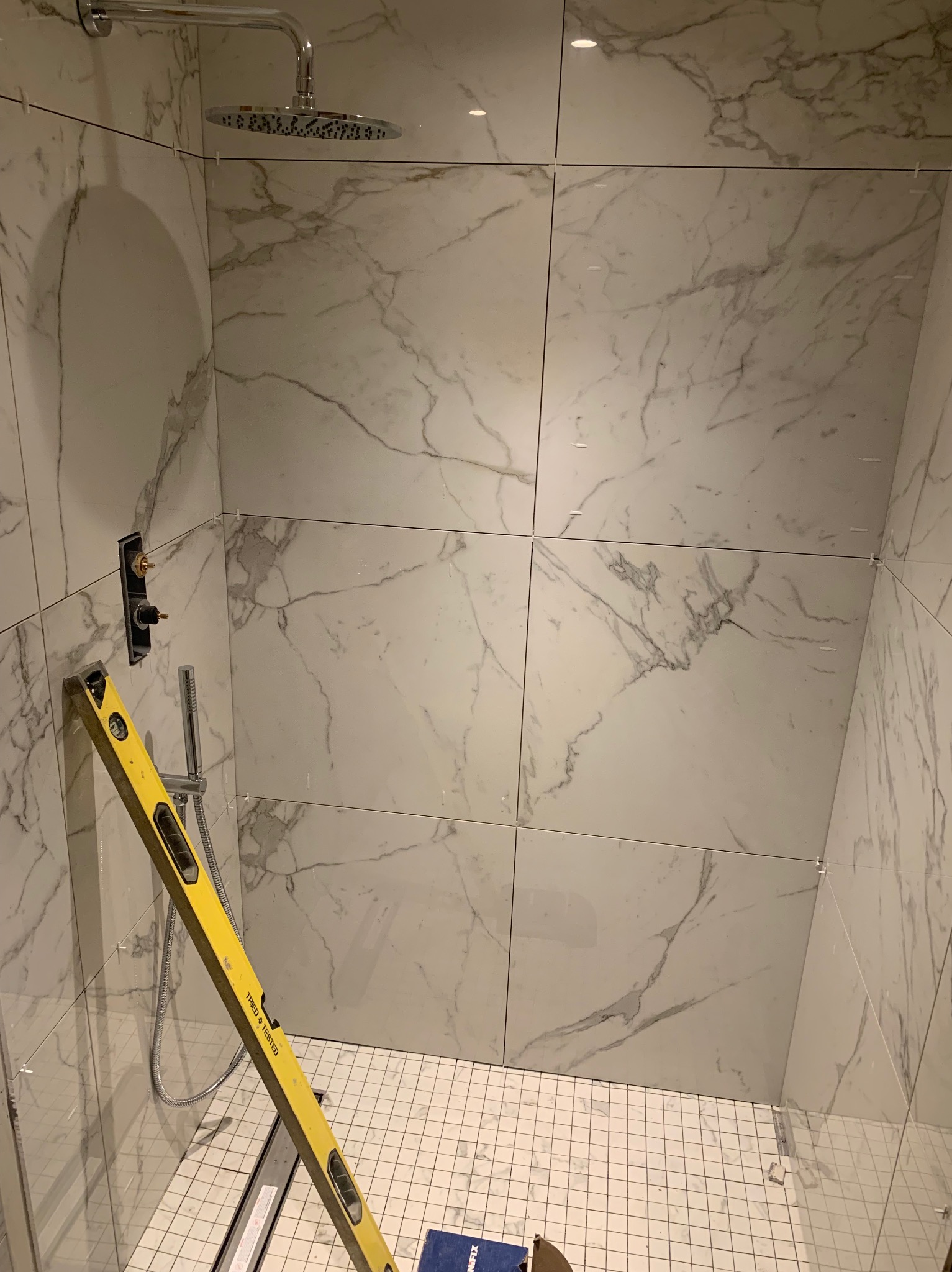 Montaż płytek imitujących marmur w kabinie prysznicowej z poziomnicą przytwierdzoną do ściany, widoczny odpływ liniowy i słuchawka prysznicowa.