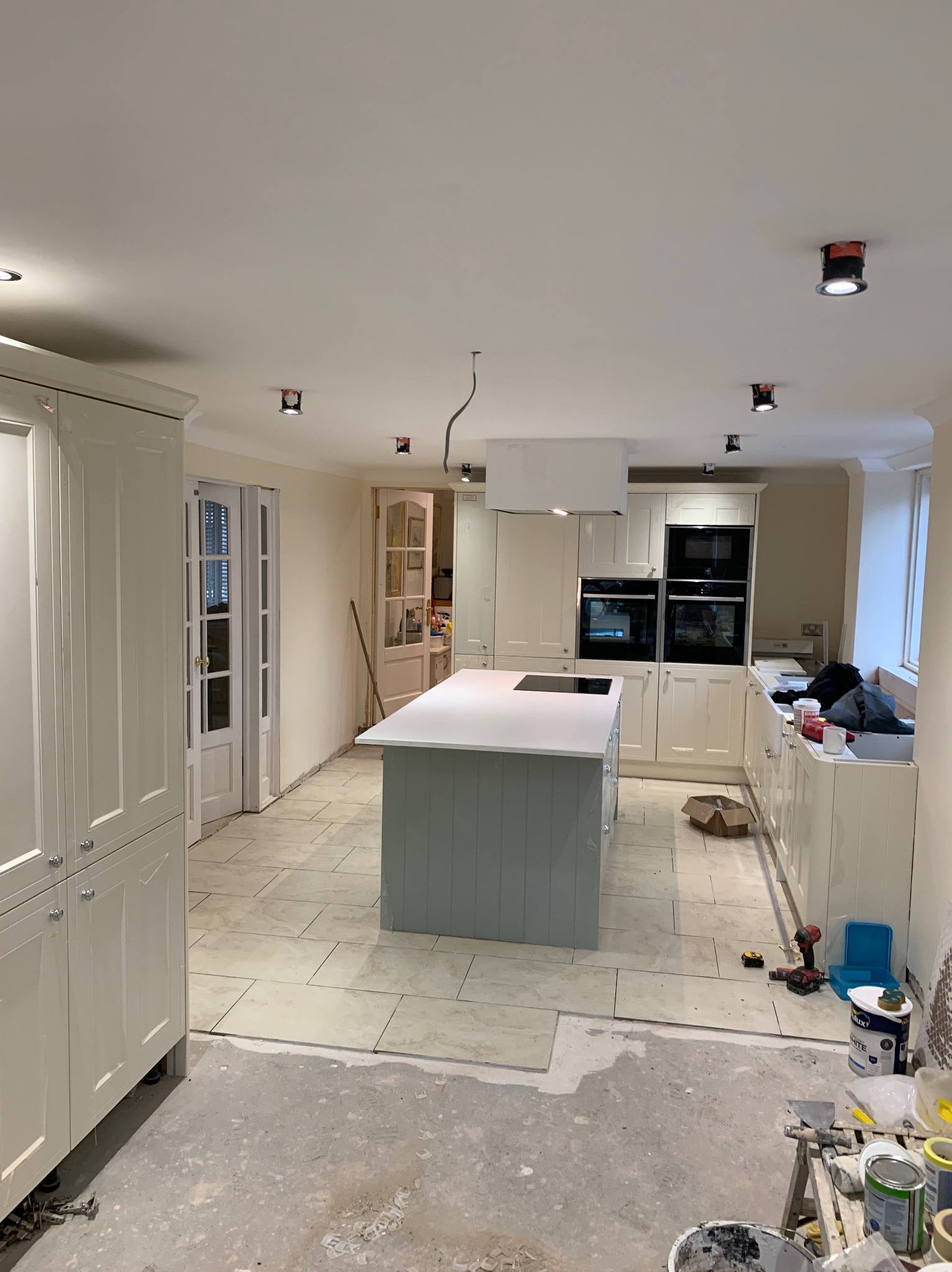 Wnętrze remontowanej kuchni z wyspą, widoczne niedokończone prace glazurnicze na podłodze, narzędzia i materiały malarskie, zabudowa kuchenna w stylu klasycznym z białymi frontami i czarnymi...