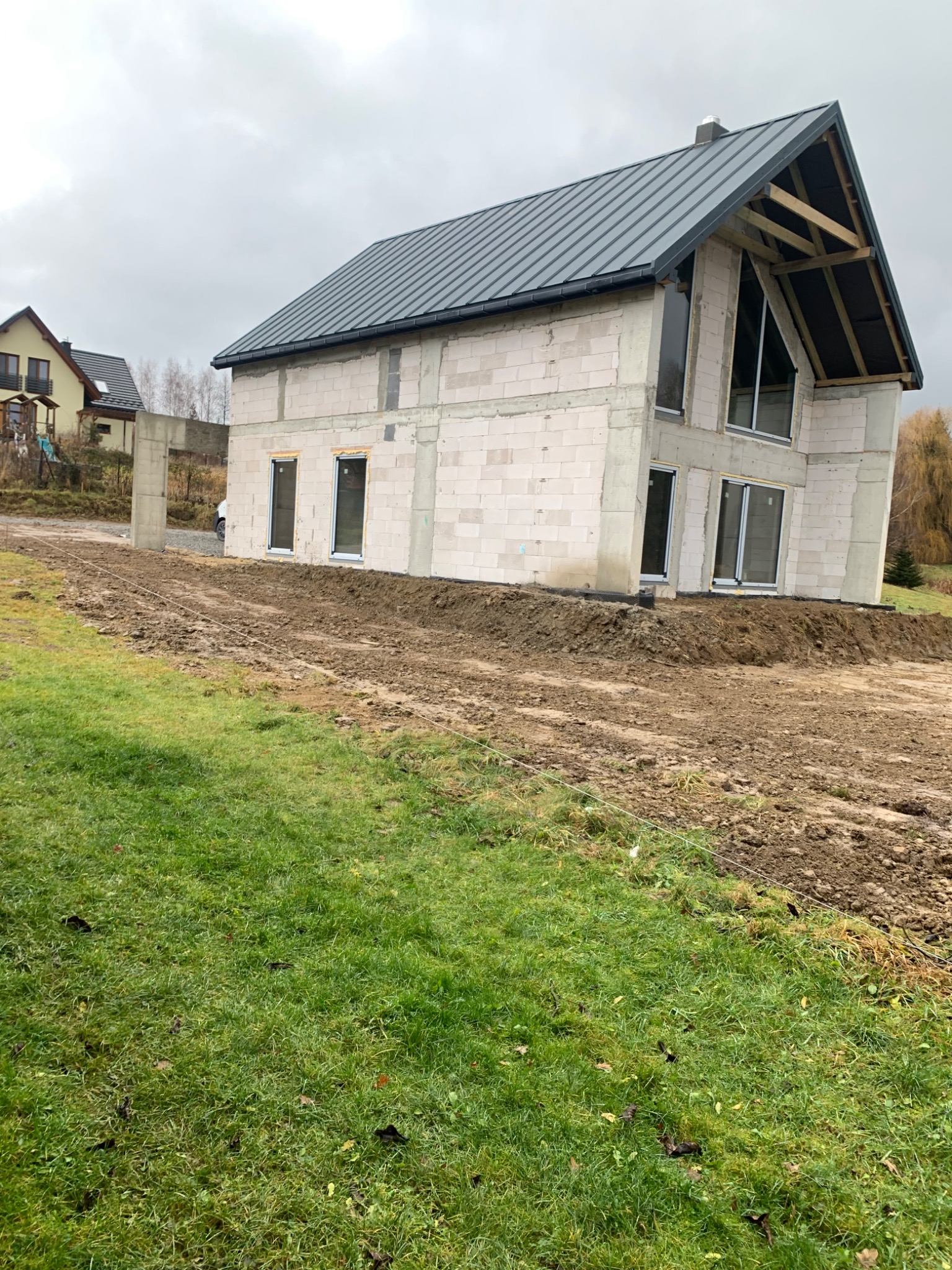 Surowy stan domu z białych bloczków z zamontowanymi oknami i szarą blachą na dachu, widoczne drewniane krokwie szczytu, na nierównym terenie z trawą i ziemią, w tle inne domy.