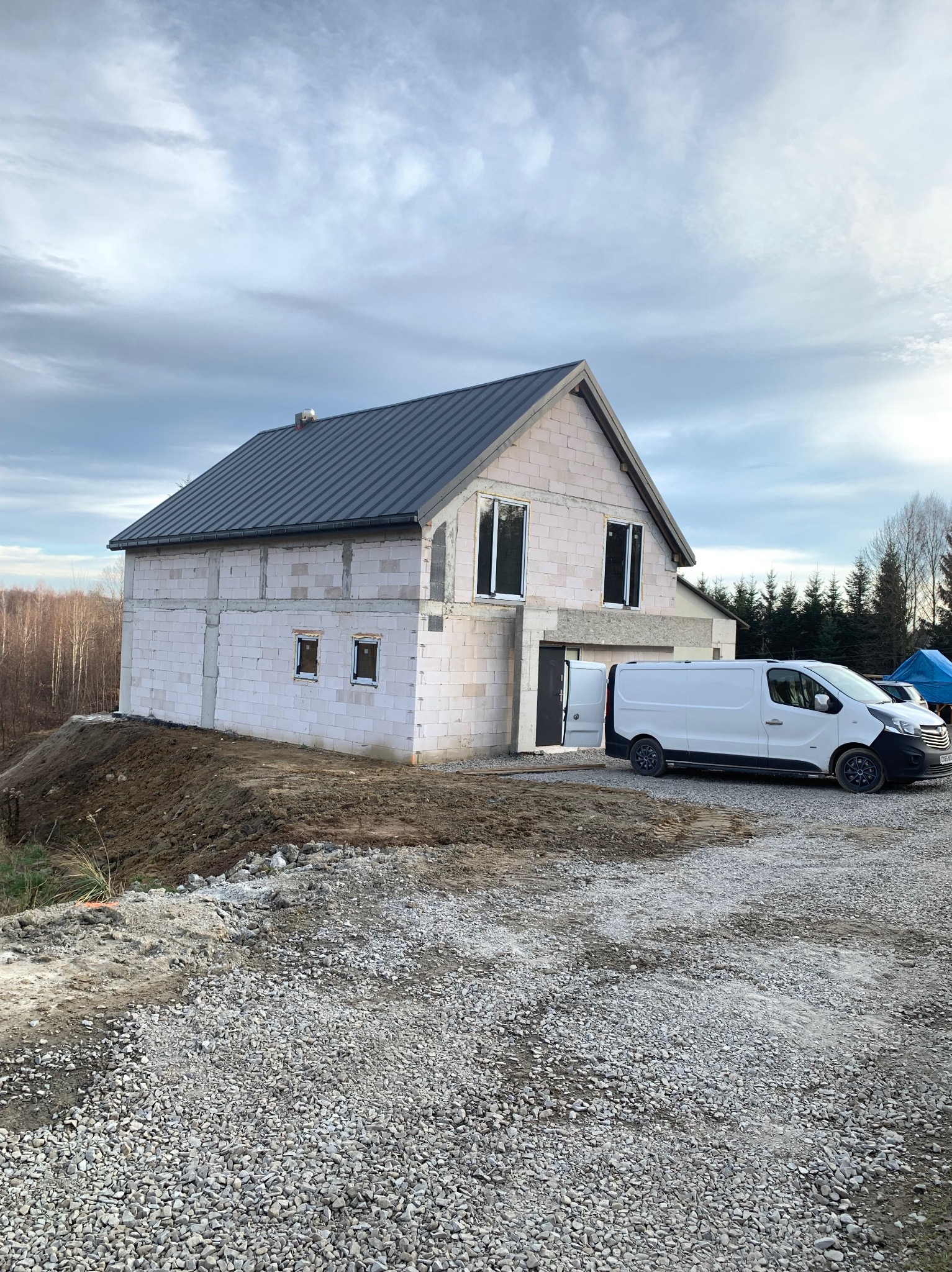 Surowy stan domu murowanego z pustaków, z zamontowaną stolarką okienną i dachem z blachy, obok zaparkowany biały bus dostawczy