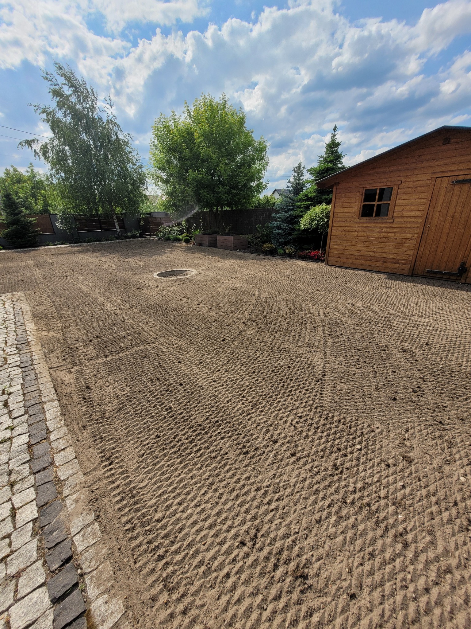 Przygotowany teren pod trawnik z widocznymi śladami po bronowaniu, obrzeże z kostki granitowej, drewniana szopa w tle, błękitne niebo z chmurami.