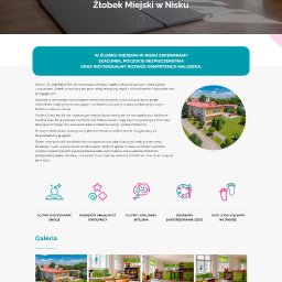 RYCHLAK.DESIGN ROBERT RYCHLAK - Sala żłobka w jasnych barwach z drewnianymi meblami i kolorowymi zabawkami. Na podłodze szare maty piankowe. Widoczne okna z dekoracjami dziecięcymi.