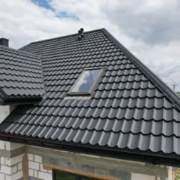 Nowy, ciemnoszary dach z blachodachówki modułowej z oknem dachowym na surowym budynku z betonu komórkowego w słoneczny dzień.