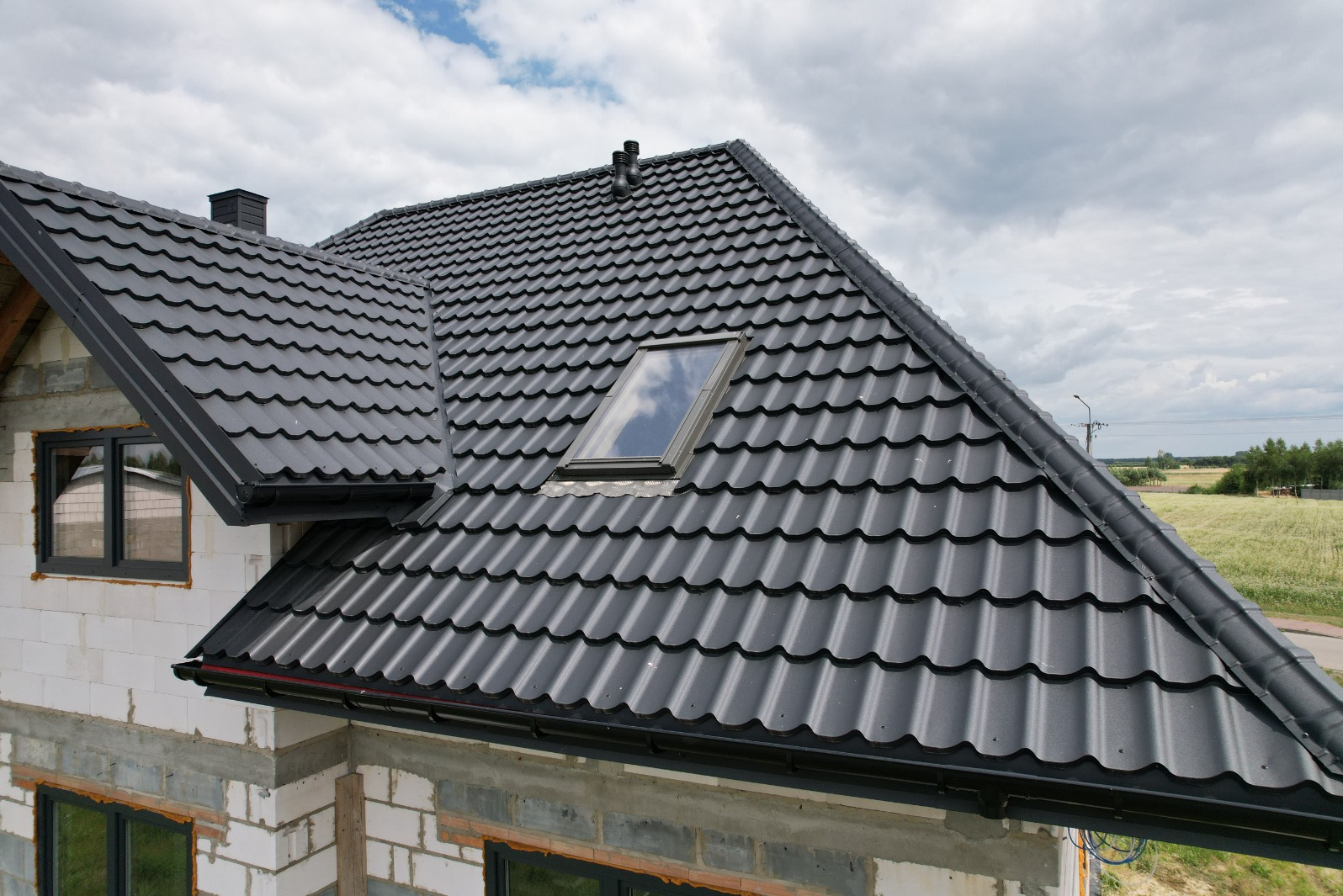 Nowy, ciemnoszary dach z blachodachówki modułowej z oknem dachowym na surowym budynku z betonu komórkowego w słoneczny dzień.