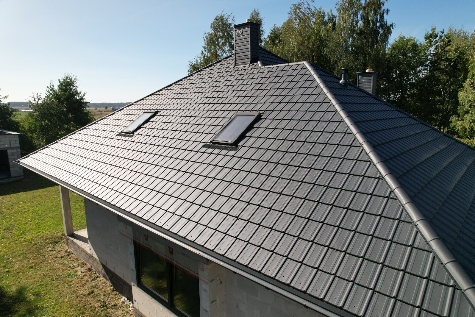 Nowy, grafitowy dach z blachy na domu w stanie surowym otwartym, z dwoma oknami dachowymi i kominem, widok z góry na tle zieleni.