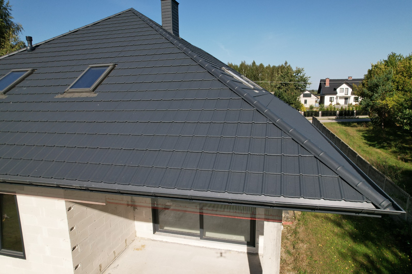 Nowy, grafitowy dach z blachy trapezowej na domu w trakcie budowy, widoczne okna dachowe i orynnowanie, w tle dom jednorodzinny i zieleń.