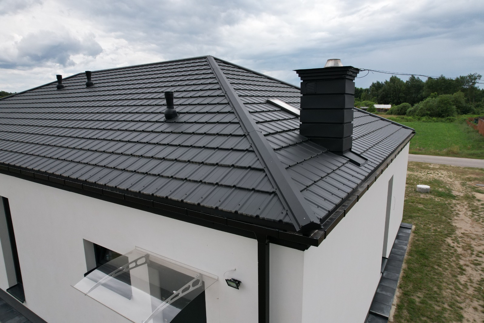 Nowoczesny dom z ciemnoszarą blachą na dachu, widoczne kominy wentylacyjne i system rynnowy w kolorze dachu, biała elewacja i przezroczysty daszek nad wejściem.