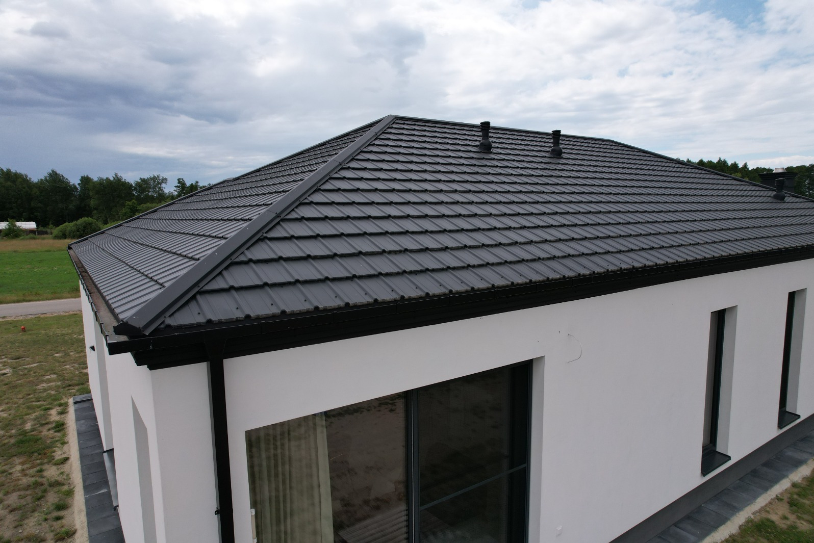 Czarny dach z blachy trapezowej na nowoczesnym, białym domu jednorodzinnym, widok z góry, z widocznymi rynnami i kominami.