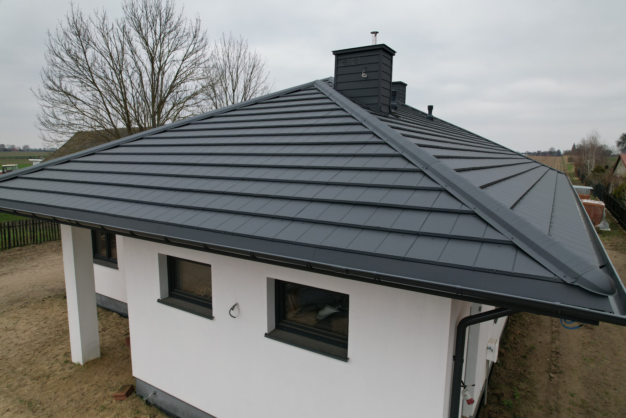 Nowoczesny, grafitowy dach z blachy na białym domu jednorodzinnym, widok z góry, kominy w kolorze dachu, jesienne drzewo w tle.