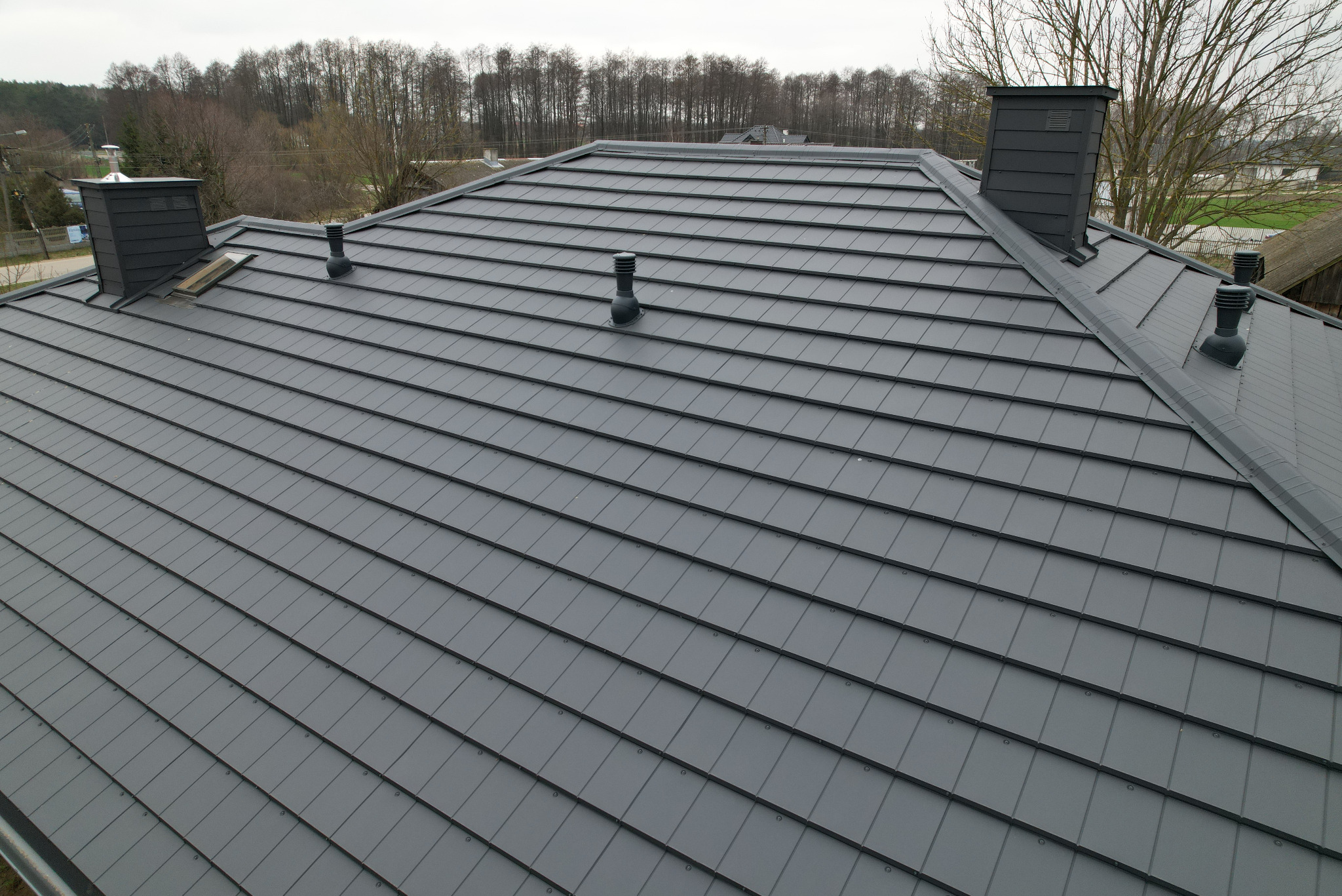Nowoczesny dach z szarej blachy modułowej z kominami i odpowietrznikami, widok z góry na tle drzew.