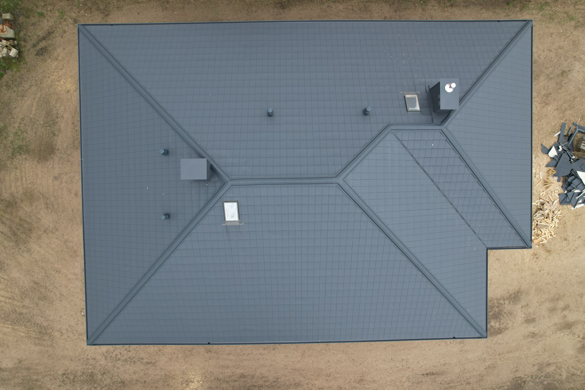 Widok z góry na nowo położony, grafitowy dach z dachówki modułowej na domu jednorodzinnym, z widocznymi kominami, oknami dachowymi i pozostałościami materiałów budowlanych na ziemi.