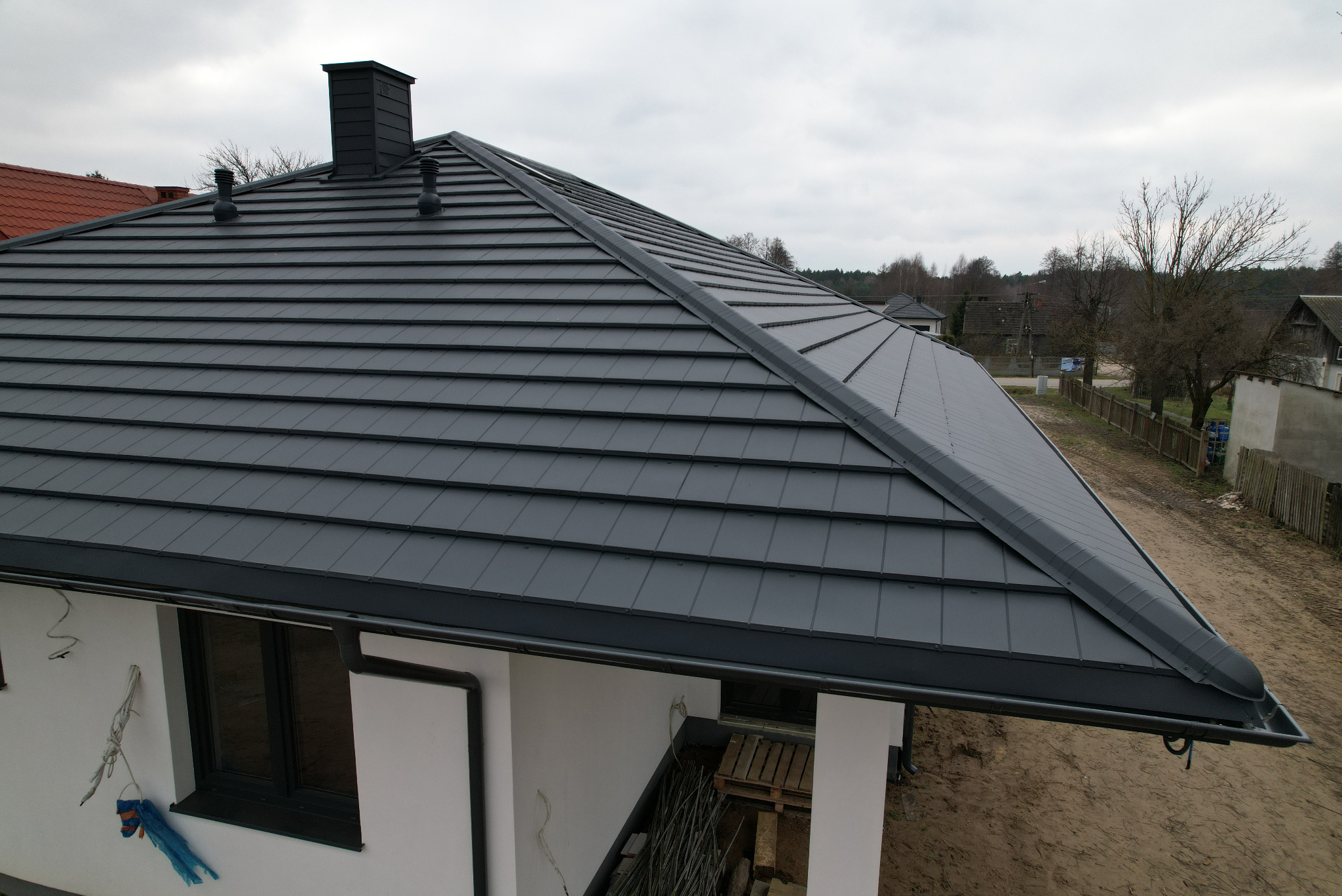 Nowoczesny dach z grafitowej blachy modułowej z kominem i wentylacją, widok z góry, na tle zabudowy jednorodzinnej i pochmurnego nieba.