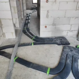 Surowe wnętrze w trakcie budowy z widocznymi przewodami elektrycznymi ułożonymi w peszlach i przymocowanymi do podłoża zielonymi opaskami oraz przewodami prowadzonymi po ścianie.