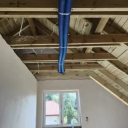 Dwie niebieskie rury wentylacyjne przymocowane do drewnianej konstrukcji dachu na poddaszu, widoczne przewody elektryczne i okno z widokiem na dom z czerwoną dachówką.