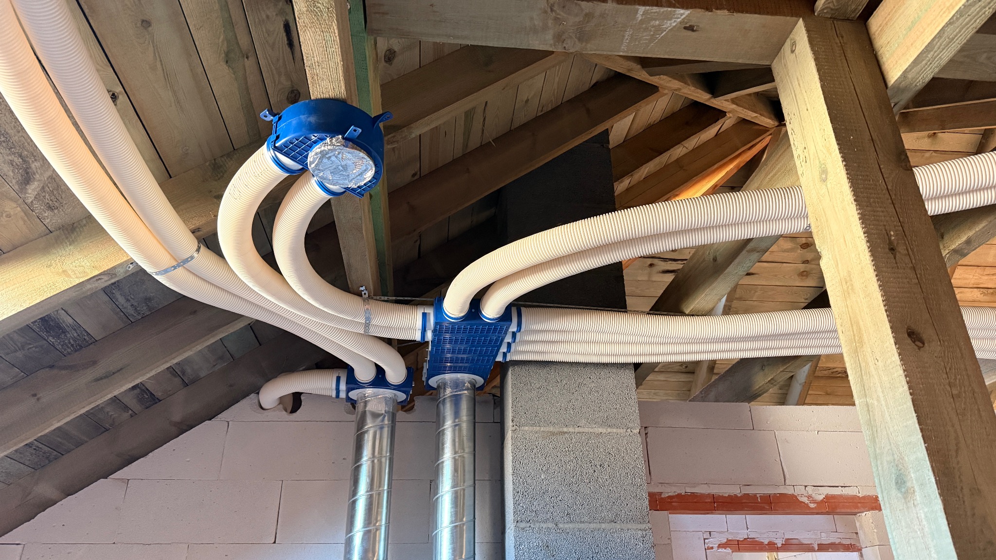 Rozbudowana instalacja rekuperacji z białymi, karbowanymi rurami i niebieskimi elementami łączeniowymi, zamontowana na poddaszu z drewnianą konstrukcją dachu.