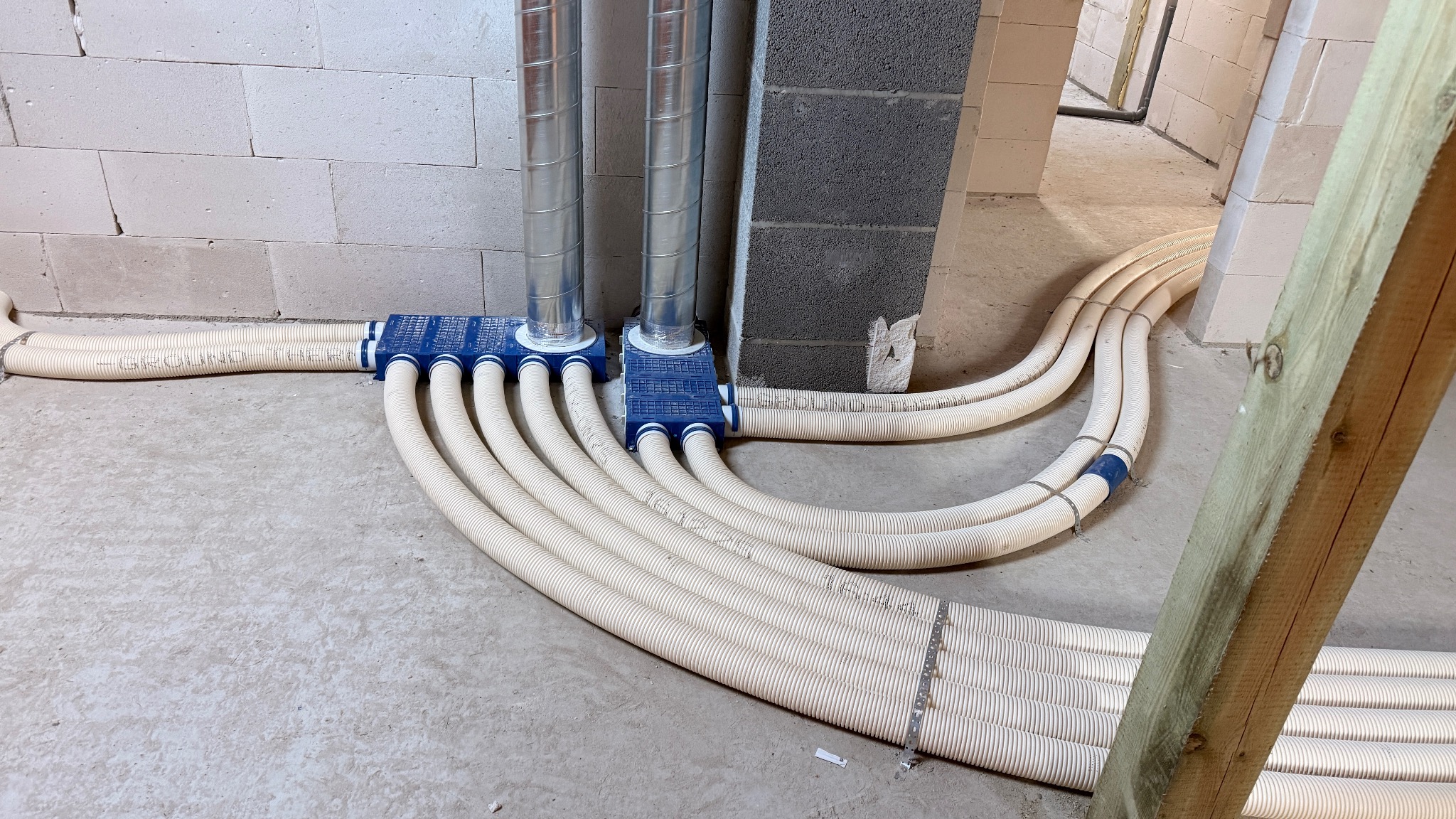 Rozprowadzenie rur wentylacyjnych w surowym wnętrzu. Widoczne metalowe rury pionowe i elastyczne rury poziome, połączone niebieskimi rozdzielaczami.