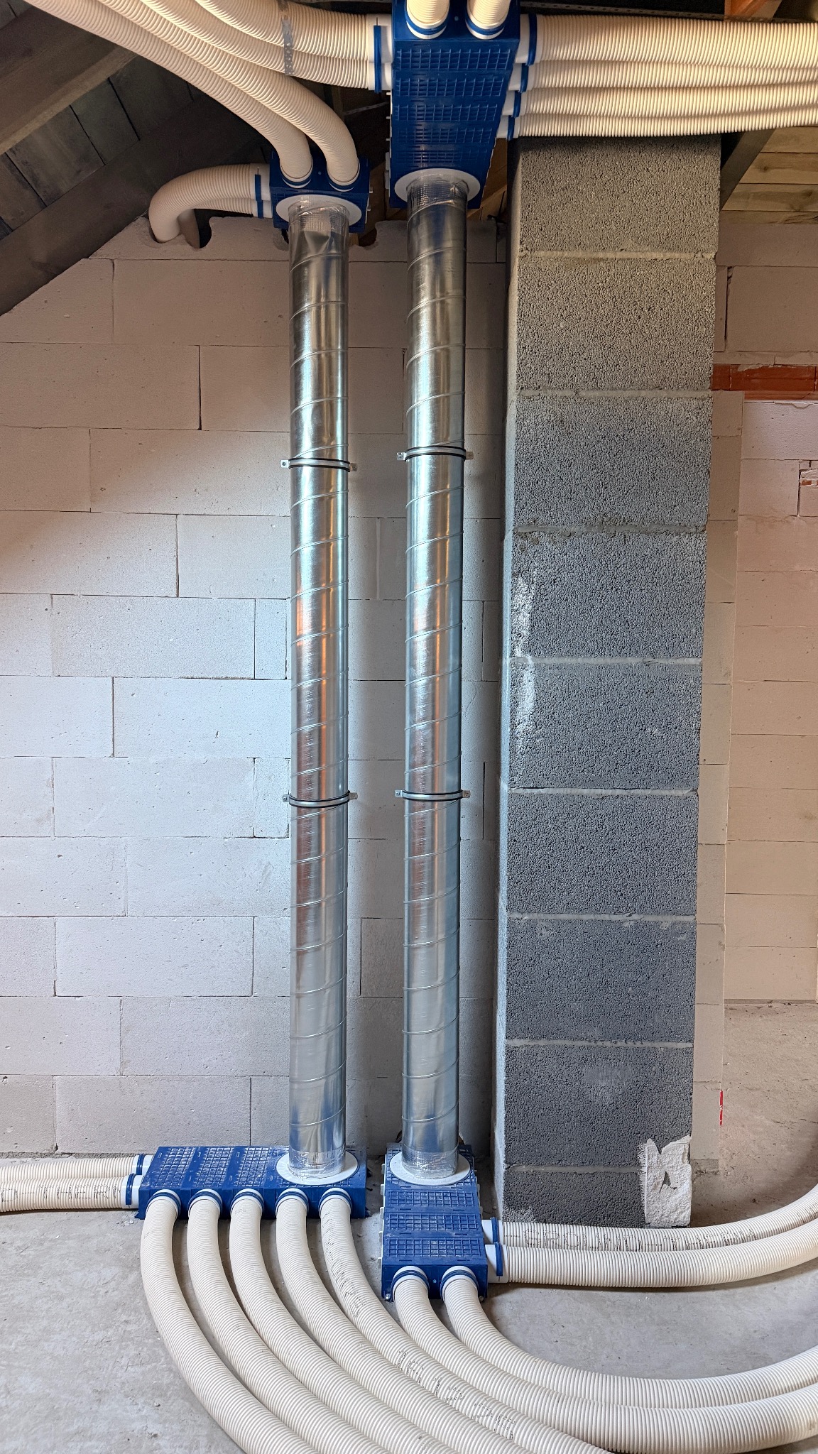 Dwie metalowe rury wentylacyjne z niebieskimi skrzynkami rozdzielczymi i białymi, elastycznymi przewodami w surowym wnętrzu budynku z bloków betonowych.