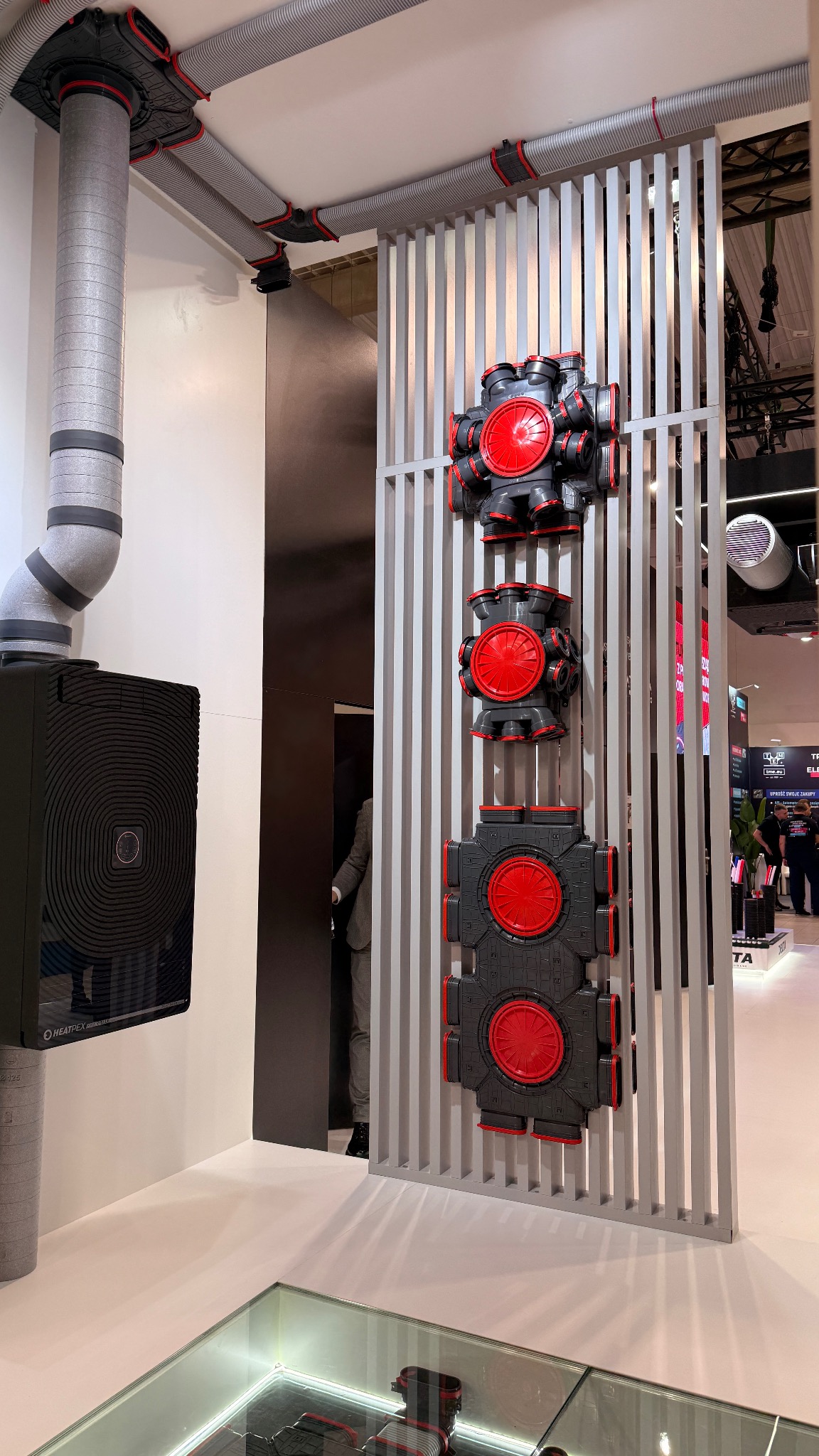 Szare rury wentylacyjne i czarno-czerwone rozdzielacze powietrza Heatpex na ekspozycji targowej, widoczne elementy instalacji rekuperacji.