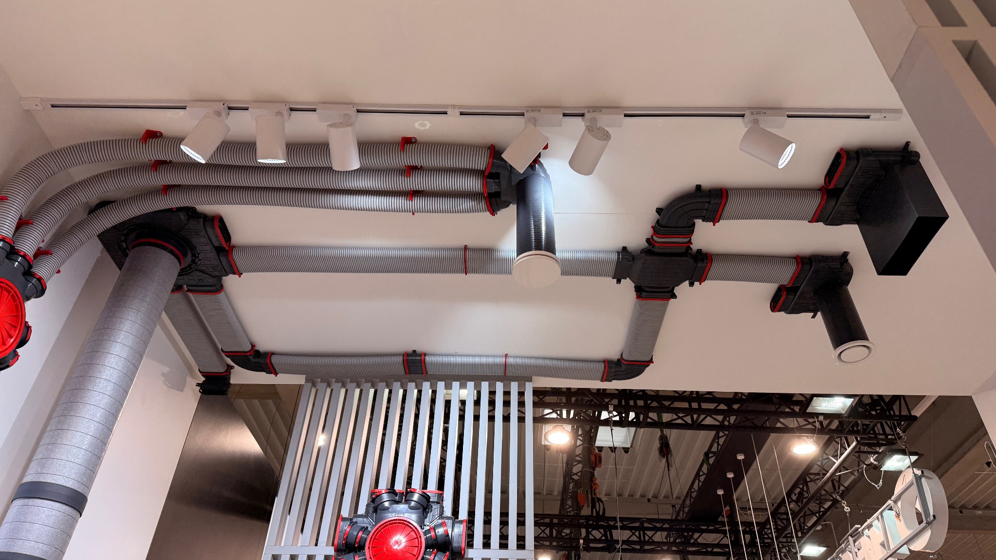 Szare, elastyczne rury wentylacyjne z czerwonymi akcentami, zamontowane pod sufitem z oświetleniem punktowym. Widok instalacji rekuperacji z perspektywy żabiej.