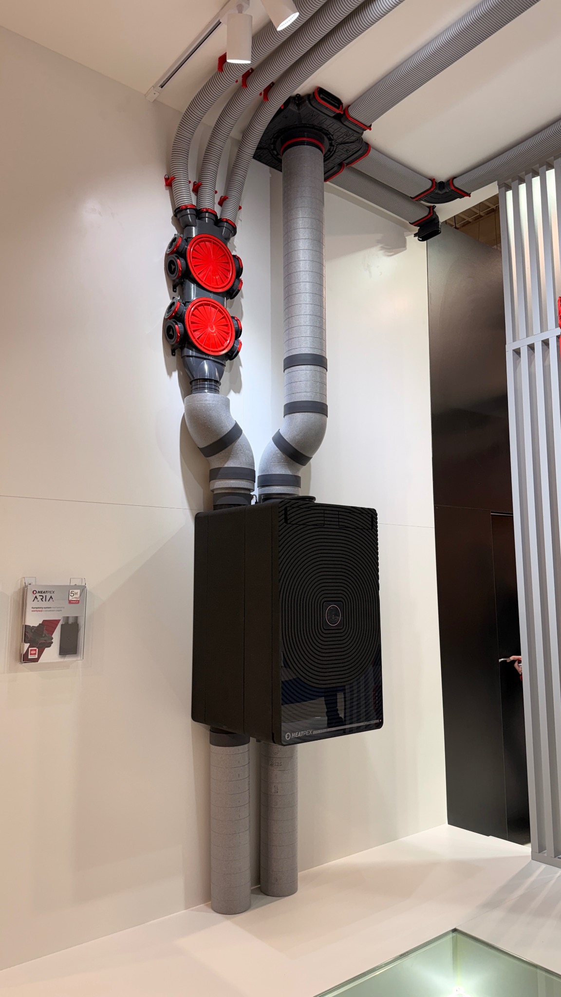Kompletny system rekuperacji Heatpex z szarymi rurami wentylacyjnymi i czarną centralą, zamontowany na białej ścianie. Widoczne elementy montażowe i rozdzielacze powietrza.