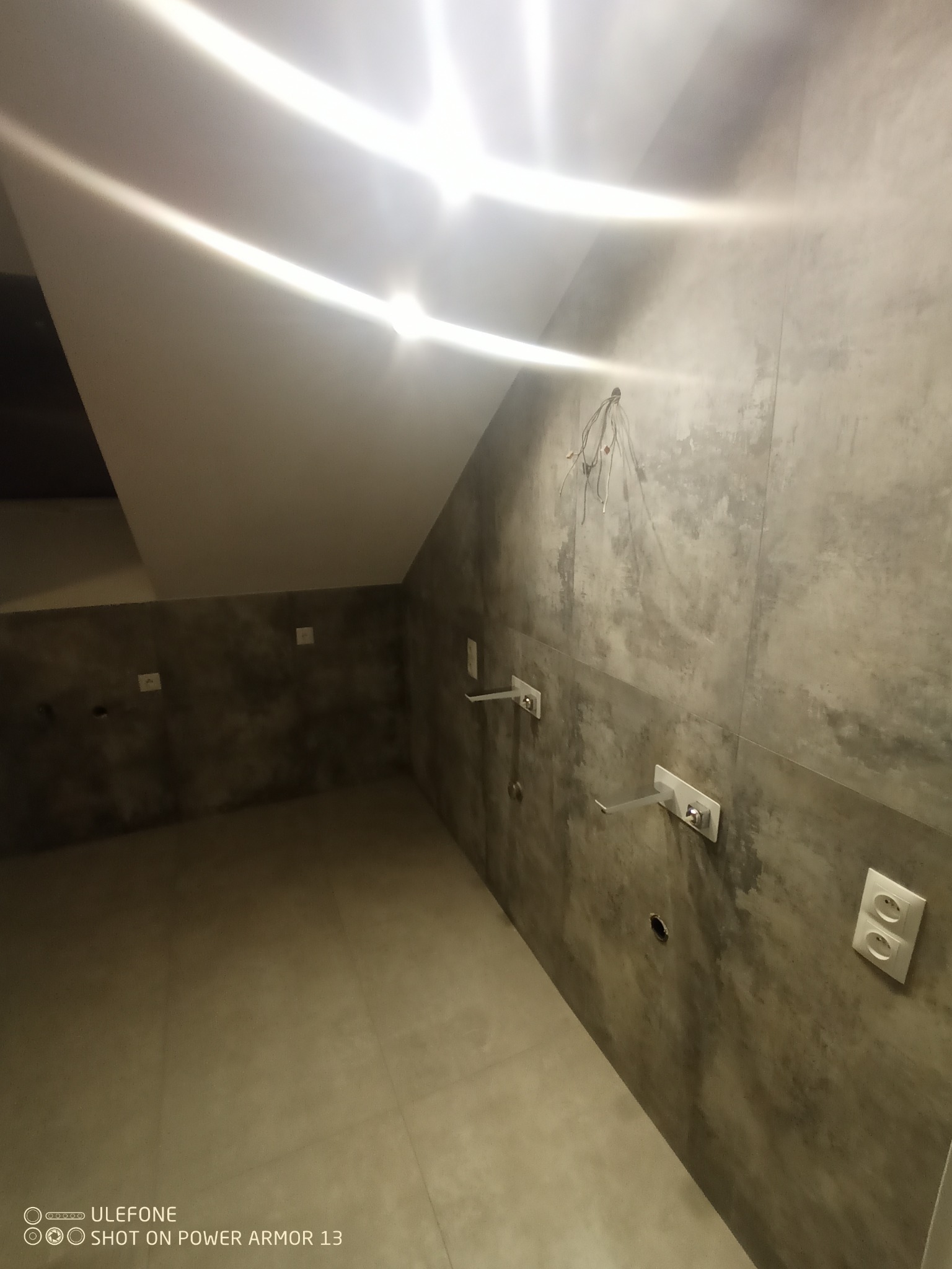 Surowa łazienka w trakcie remontu: szare płytki imitujące beton, wystające kable, przygotowane punkty hydrauliczne i gniazdka elektryczne, sufit ze skosem.