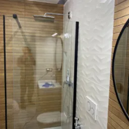 Nowoczesna łazienka z kabiną prysznicową typu walk-in, wykończona płytkami imitującymi drewno i białymi płytkami 3D w geometryczny wzór, z widoczną armaturą i okrągłym lustrem.