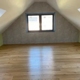 Poddasze po remoncie: drewniana podłoga z widocznymi słojami, ściana z geometryczną tapetą w odcieniach beżu i brązu, okno zabezpieczone niebieską taśmą malarską.