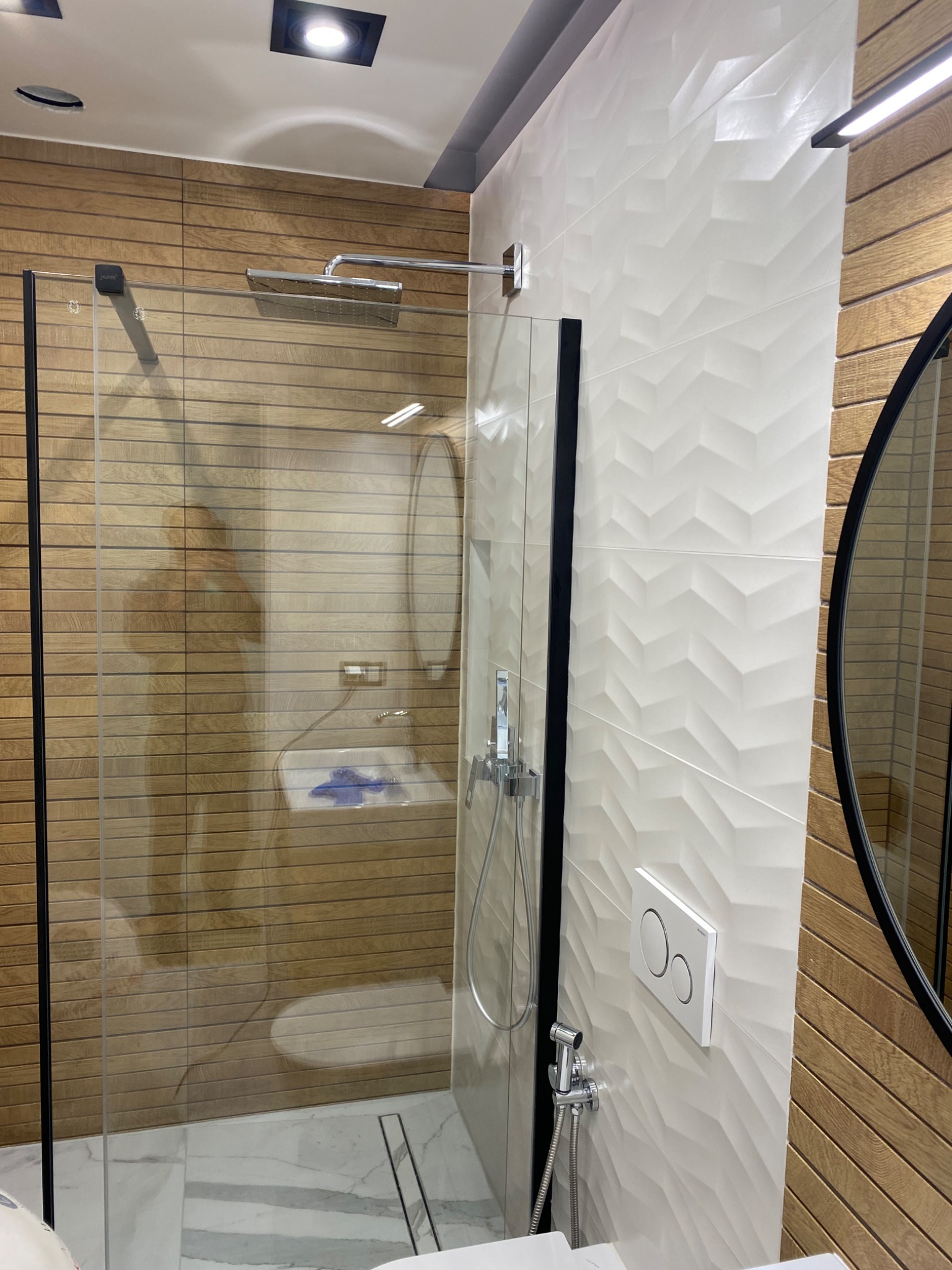 Nowoczesna łazienka z kabiną prysznicową typu walk-in, wykończona płytkami imitującymi drewno i białymi płytkami 3D w geometryczny wzór, z widoczną armaturą i okrągłym lustrem.
