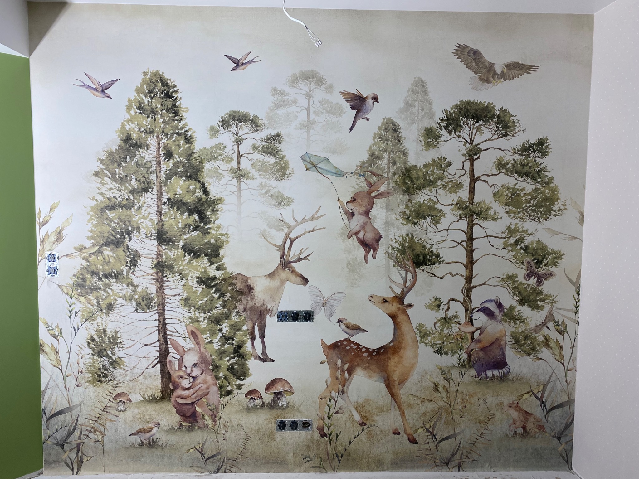 Fragment ściany z fototapetą w stylu akwareli przedstawiającą leśny krajobraz z jeleniami, królikami, ptakami i drzewami, z widocznymi gniazdkami elektrycznymi.