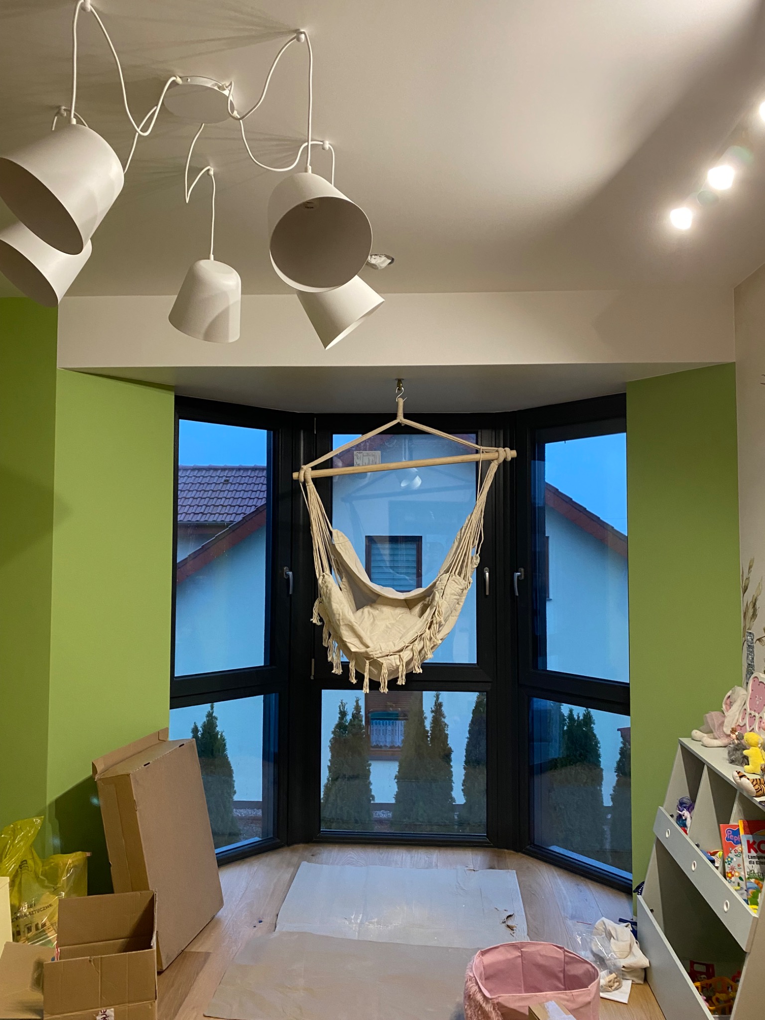 Pokój w trakcie urządzania z zieloną ścianą, beżowym hamakiem wiszącym w oknie wykuszowym i nowoczesną białą lampą sufitową.