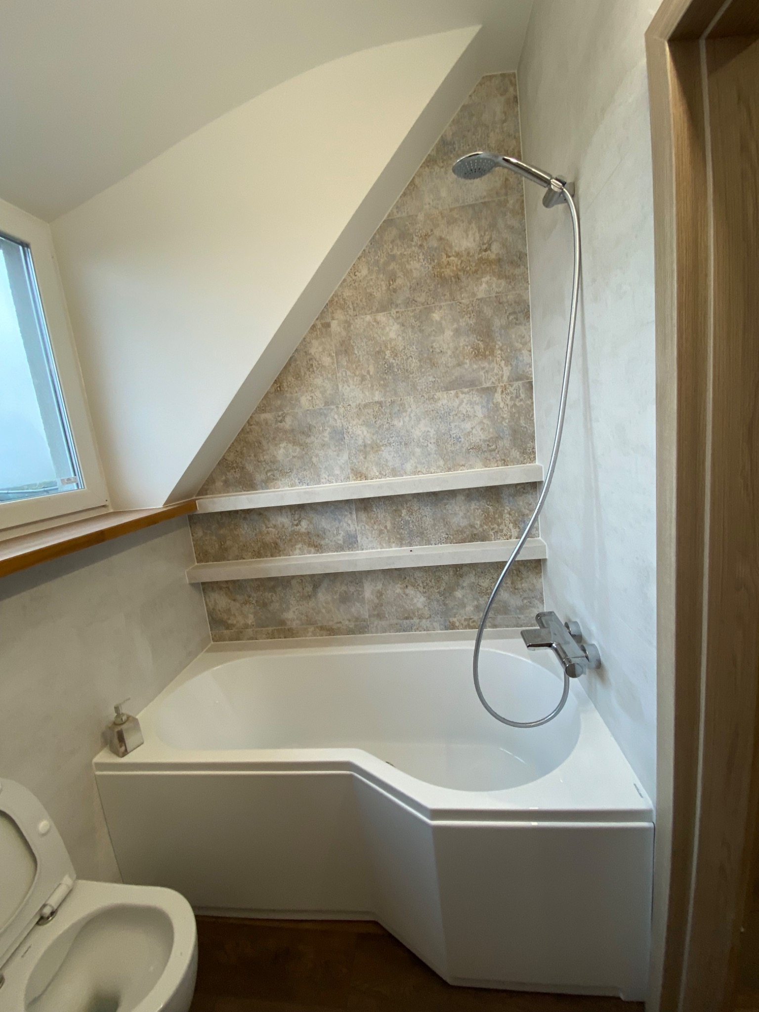 Biała wanna narożna pod skosem dachu, obok toaleta, na ścianie beżowe płytki z dwiema półkami, chromowana bateria prysznicowa.