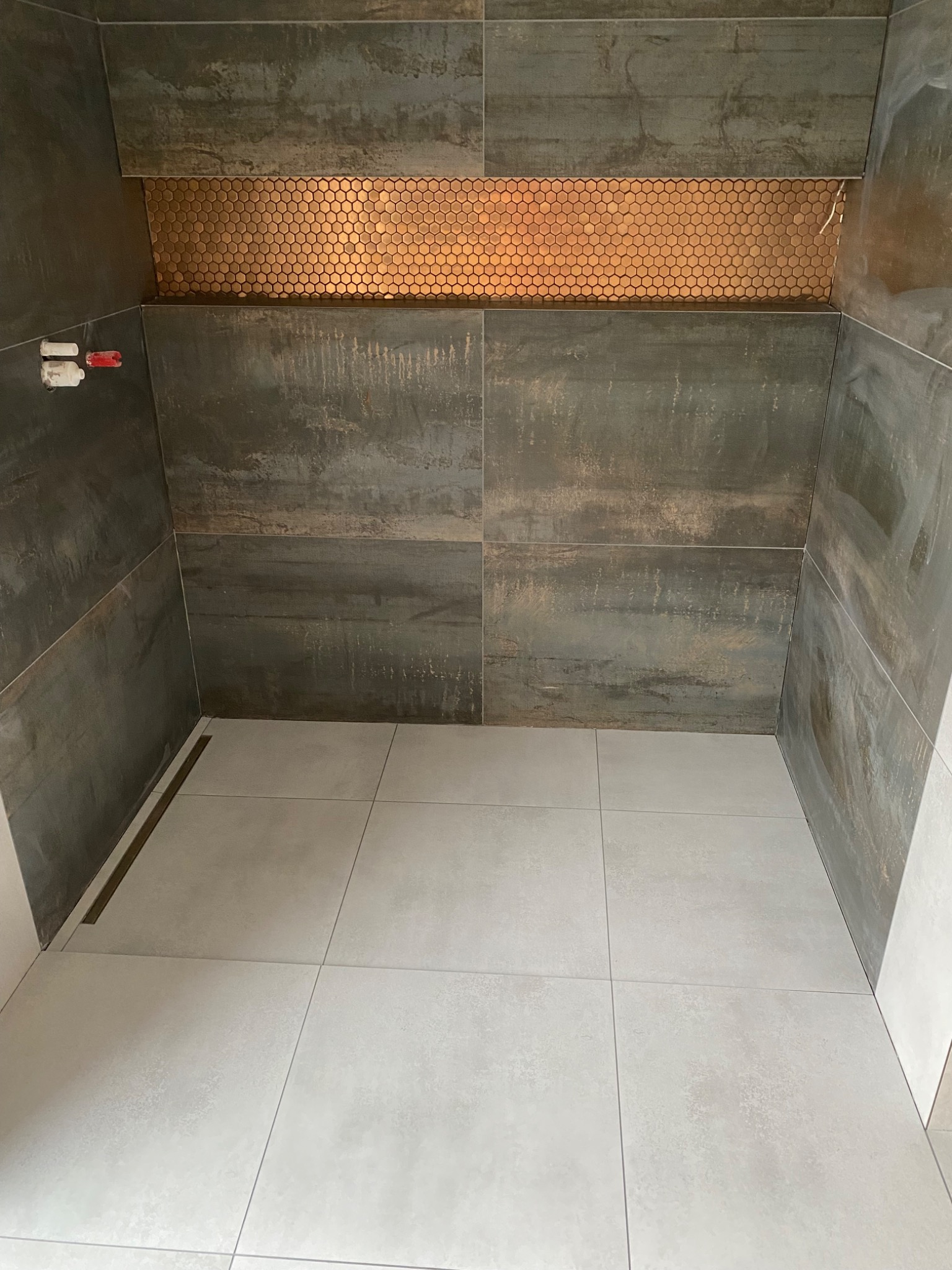 Wnętrze kabiny prysznicowej z szarymi płytkami imitującymi metal, mozaiką w odcieniach miedzi i jasnymi płytkami podłogowymi, widoczne elementy instalacji wodnej i odpływ liniowy.