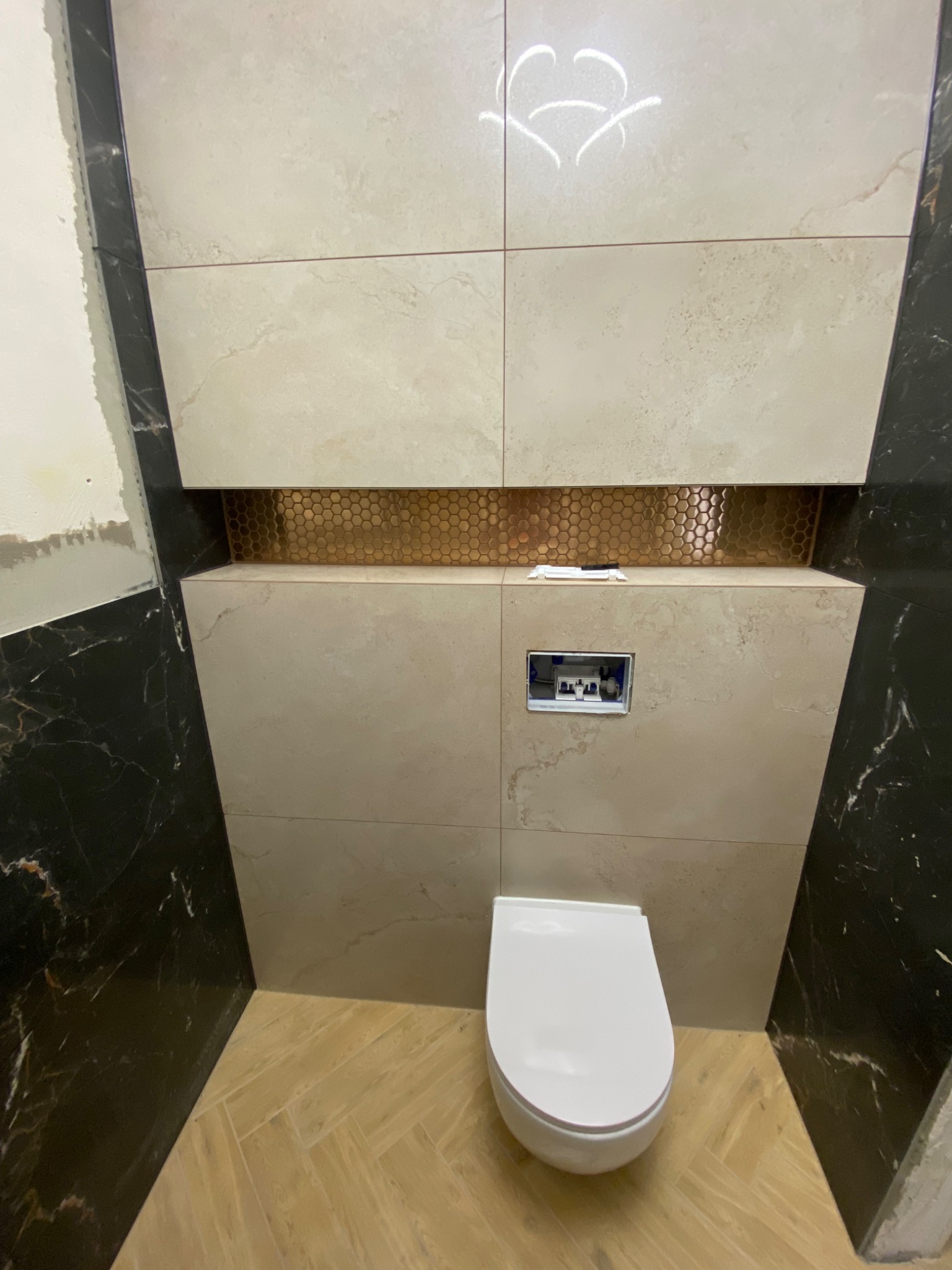 Nowoczesna łazienka z podwieszaną toaletą, beżowe płytki imitujące kamień, dekoracyjny pas z mozaiki w kolorze miedzi, czarne marmurowe akcenty na ścianach, oraz parkiet ułożony w jodełkę na podłodze.