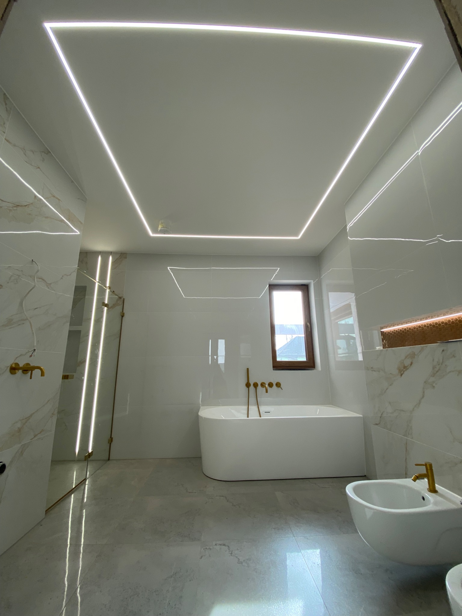 Nowoczesna łazienka z białymi marmurkowymi płytkami, podświetleniem LED w kształcie kwadratu na suficie, złotą armaturą i wolnostojącą wanną.