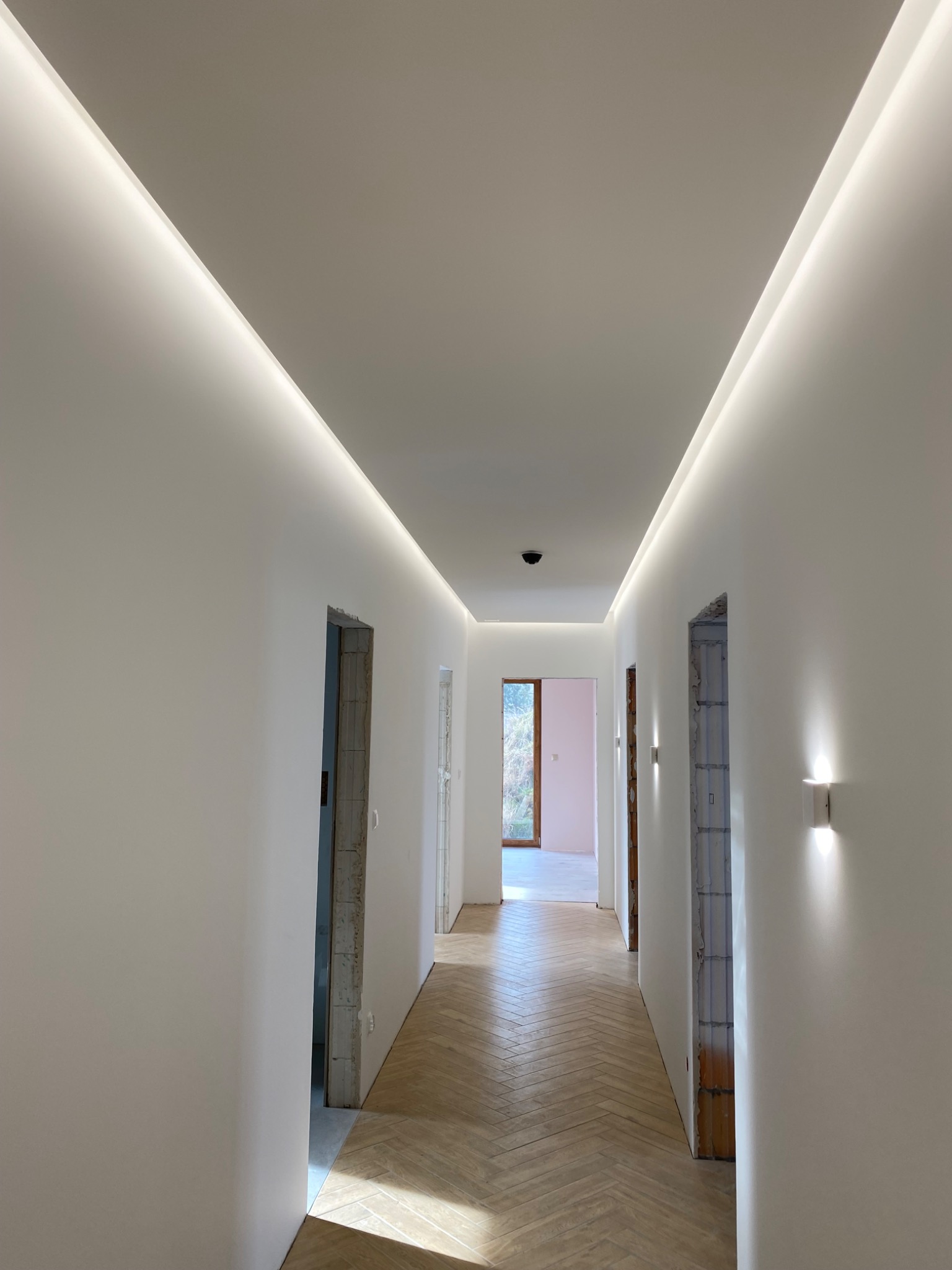 Korytarz z parkietem ułożonym w jodełkę, świeżo pomalowanymi białymi ścianami i sufitem z ukrytym oświetleniem LED. Widoczne surowe ościeżnice drzwi.