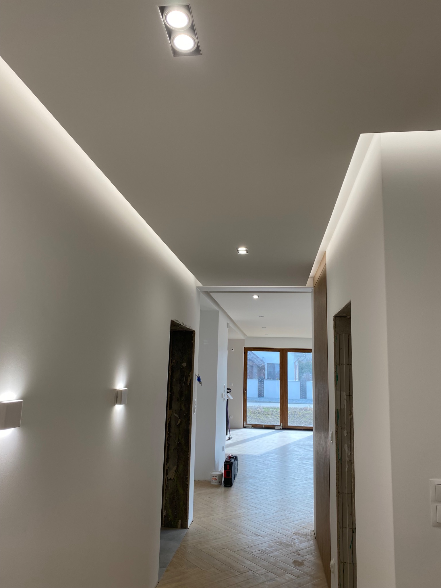 Nowoczesny korytarz z oświetleniem LED w listwach sufitowych i punktowych, parkiet ułożony w jodełkę, widoczne fragmenty ościeżnic drzwiowych w trakcie montażu.