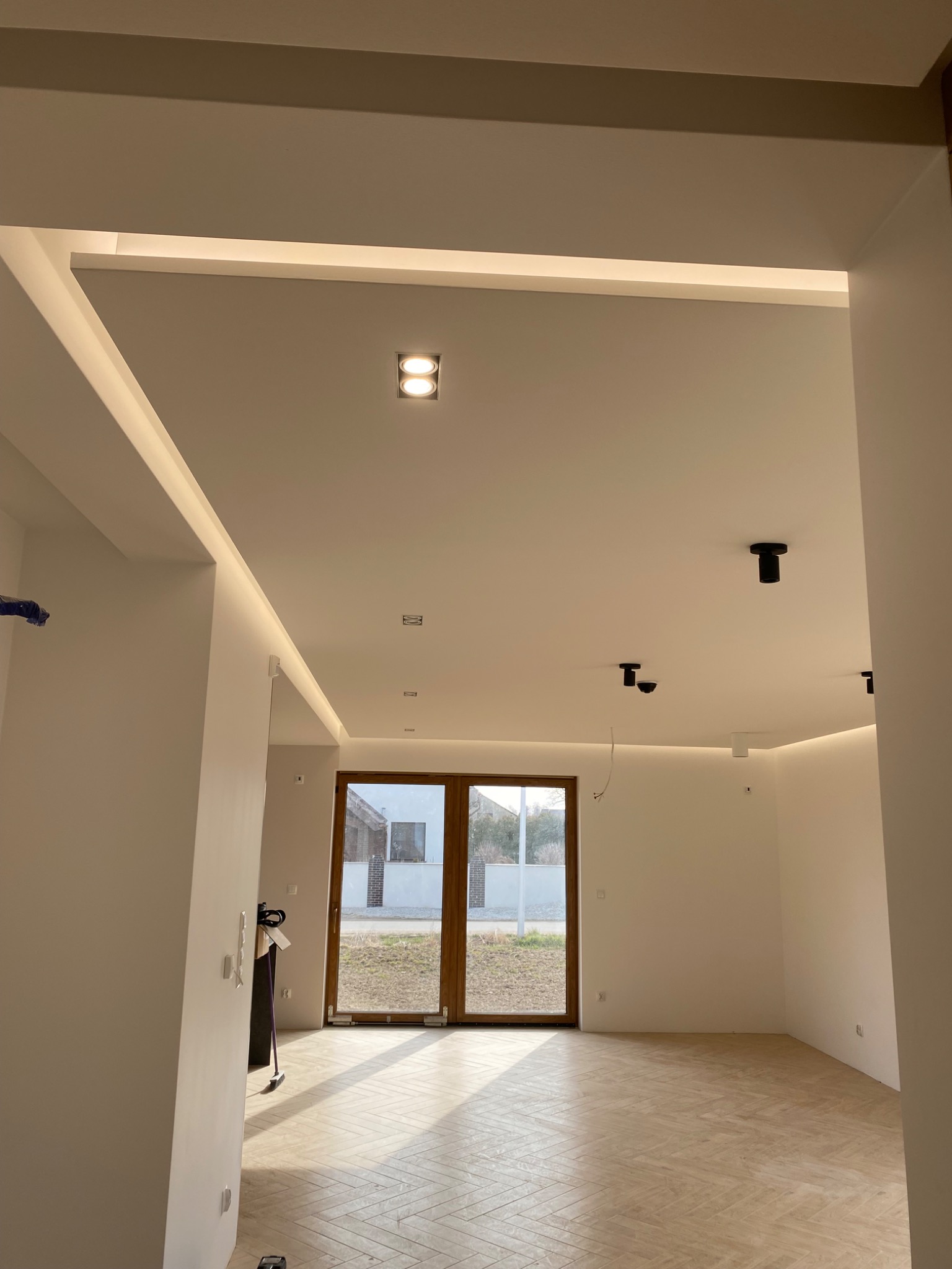 Wnętrze w stanie deweloperskim z drewnianą podłogą ułożoną w jodełkę, oświetleniem LED w suficie podwieszanym i czarnymi reflektorami punktowymi.