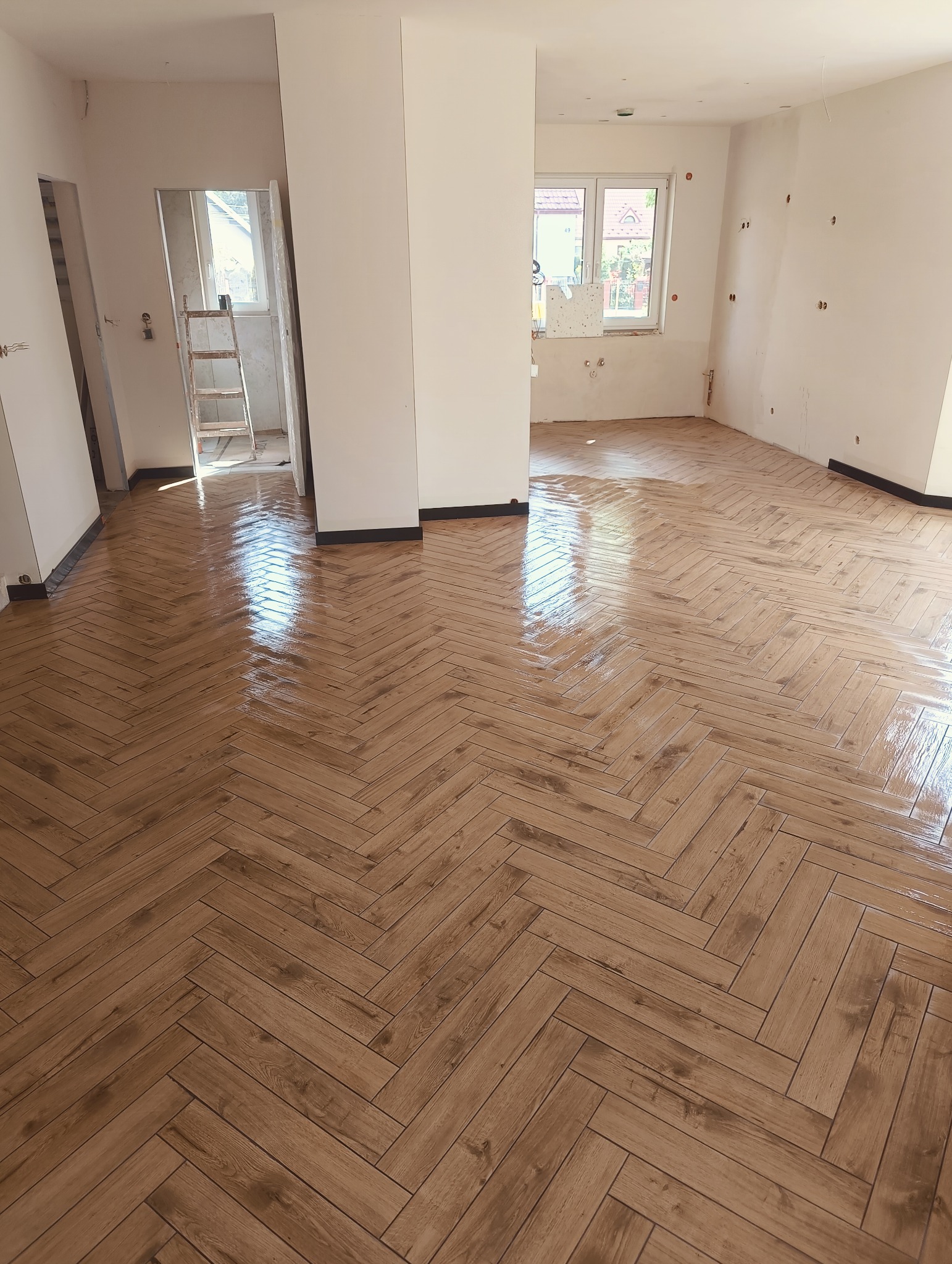 Ułożona, błyszcząca podłoga z paneli w jodełkę, widok z góry. W tle fragmenty ścian, kolumna i okno. W pomieszczeniu widoczne ślady prac wykończeniowych.