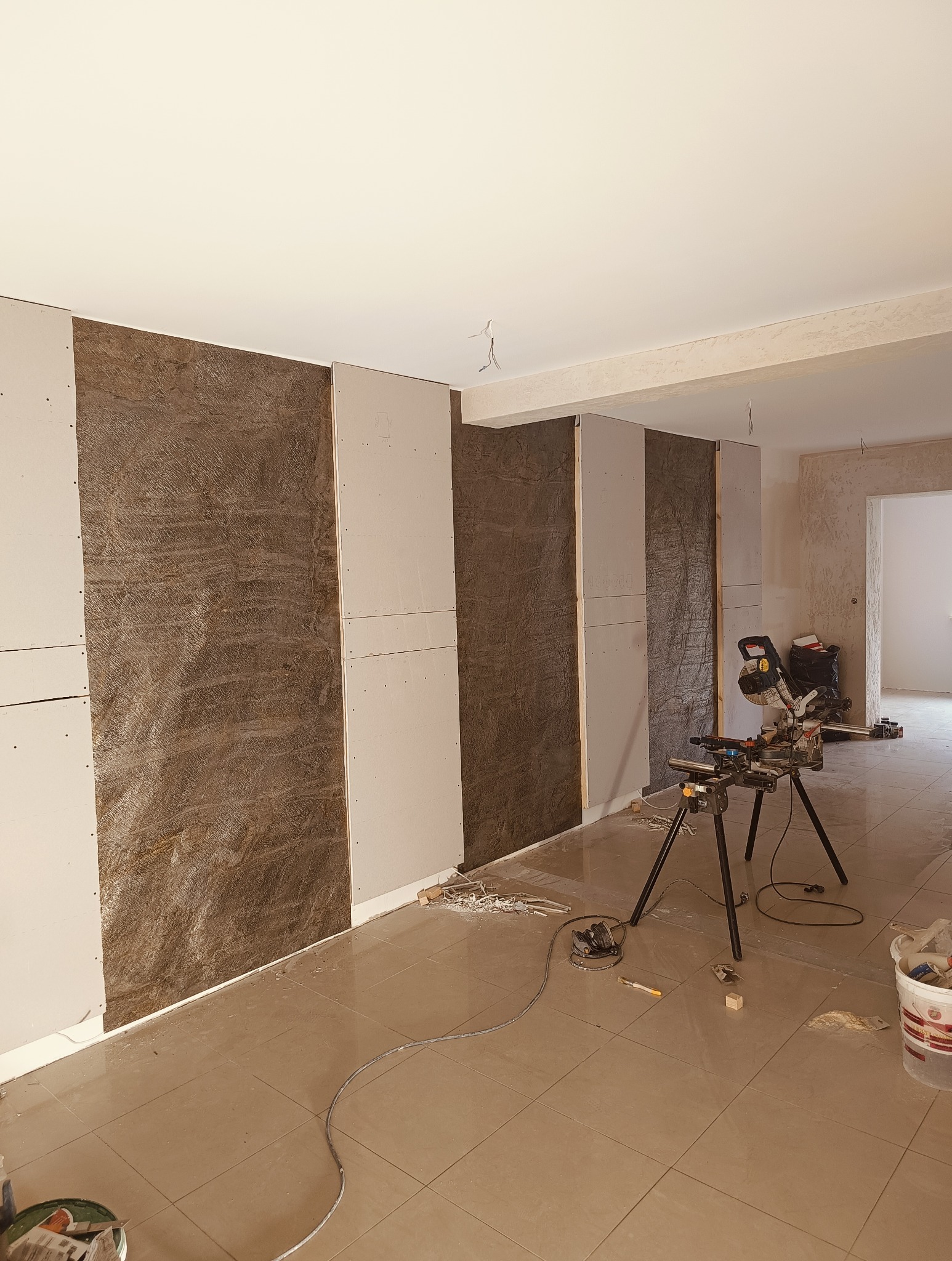 Montaż płyt karton-gips na ścianie z dekoracyjną okładziną. Widoczna piła do cięcia na statywie, podkreślająca etap prac wykończeniowych we wnętrzu.